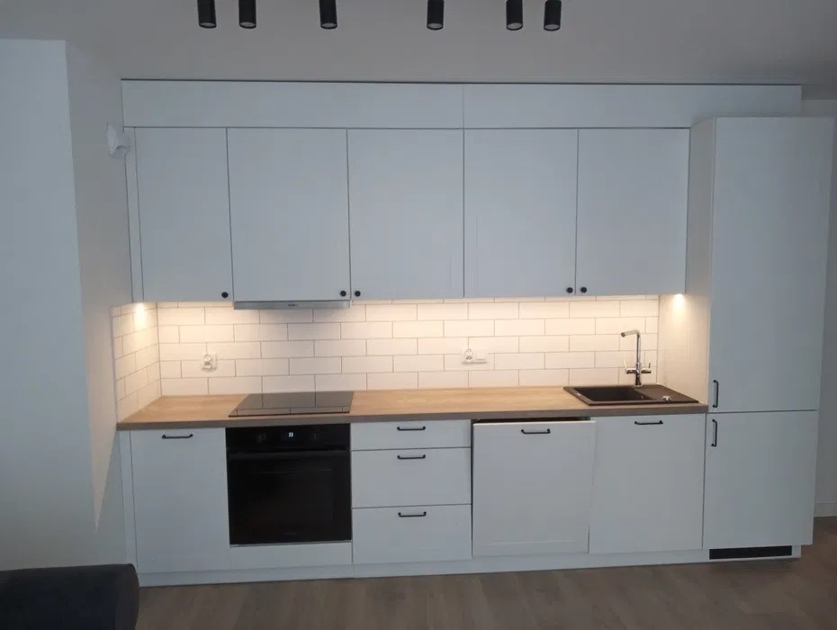 Nowoczesna, biała zabudowa kuchenna z drewnianym blatem, czarnym zlewem i płytą indukcyjną, oświetlona punktowymi lampami sufitowymi i listwą LED pod szafkami wiszącymi.