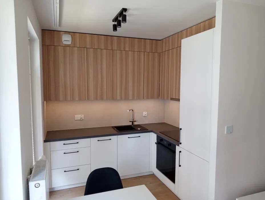 Nowoczesna kuchnia z białymi szafkami dolnymi i drewnopodobnymi górnymi, czarnym blatem i minimalistyczną armaturą, oświetlona punktowo.