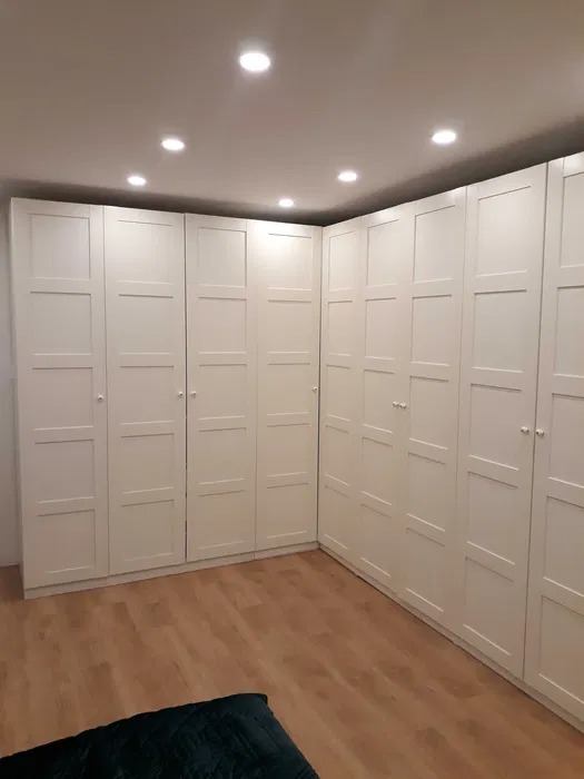 Biała, narożna zabudowa szaf z panelowymi drzwiami, oświetlona punktowymi lampami sufitowymi, na podłodze panele drewnopodobne.