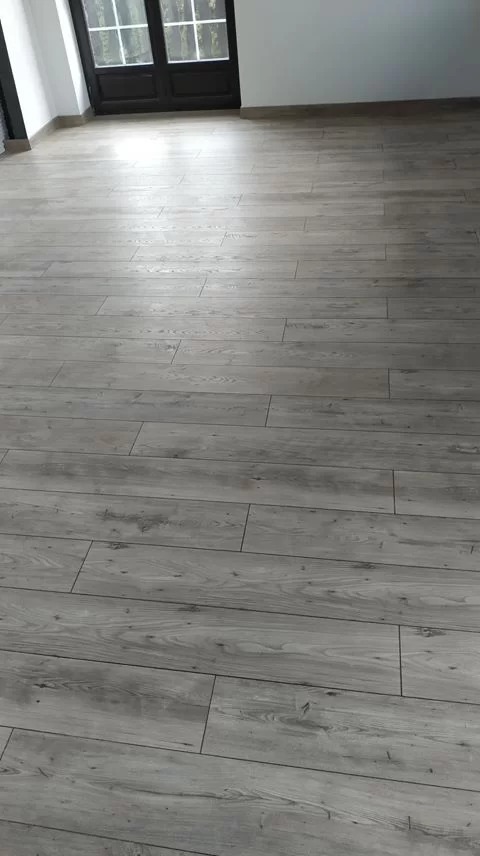 Ułożone panele podłogowe w odcieniach szarości z widoczną strukturą drewna, ciemne drzwi balkonowe w tle.