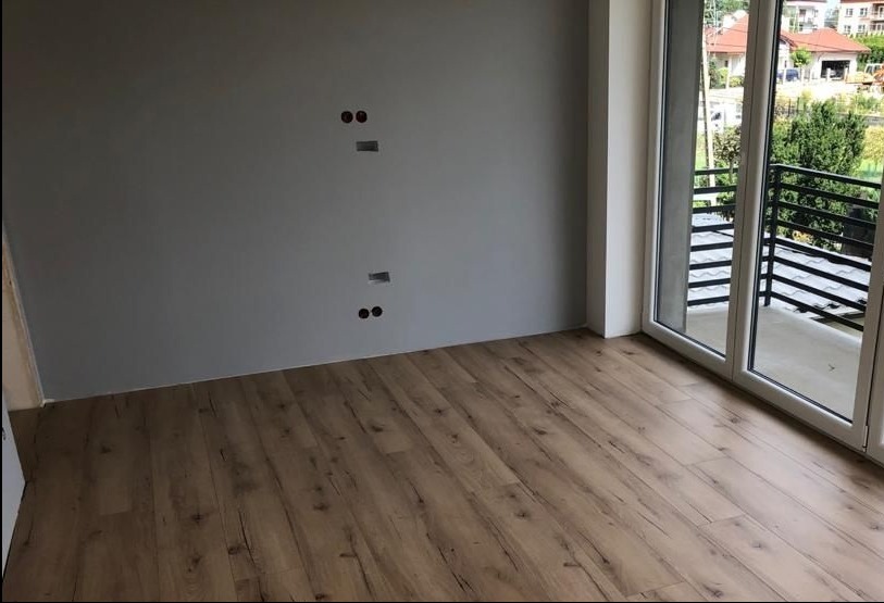 Pokój po remoncie: ułożone panele podłogowe w odcieniach brązu, szara ściana z widocznymi otworami na gniazdka elektryczne, przesuwne drzwi balkonowe z widokiem na zieleń.
