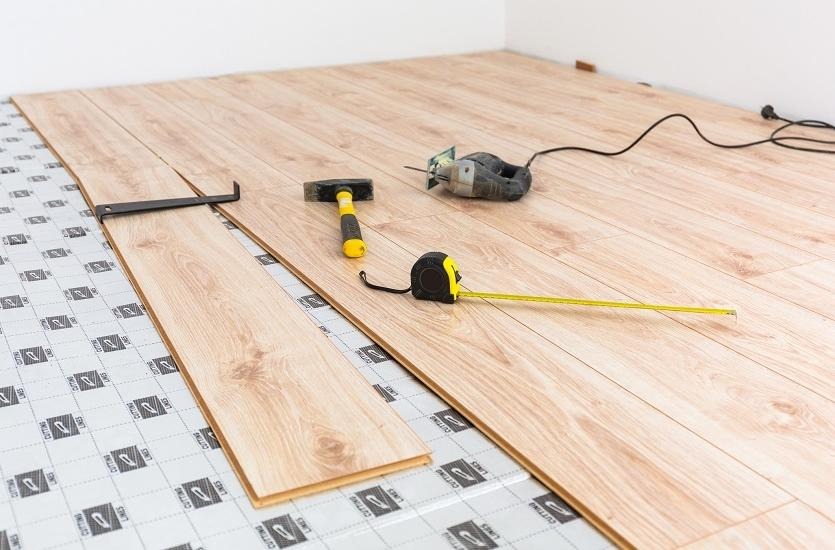 Proces układania paneli podłogowych: widoczne narzędzia, podkład z nadrukiem i fragment ułożonej podłogi z jasnego drewna.