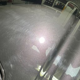 Brand New Car - Powierzchnia lakieru samochodu z widocznymi kolistymi zarysowaniami, odbijająca oświetlenie warsztatowe przed procesem polerowania.