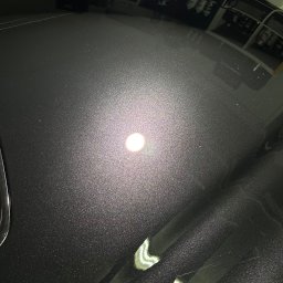 Brand New Car - Błyszcząca, ciemnoszara powierzchnia karoserii samochodu po polerowaniu, odbijająca światło lampy warsztatowej, z widocznymi refleksami i minimalnymi niedoskonałościami lakieru.