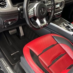 Brand New Car - Wnętrze luksusowego samochodu marki Mercedes-Benz AMG z czerwoną perforowaną skórzaną tapicerką fotela kierowcy, czarną kierownicą i dywanikami z logo AMG, widok z boku.