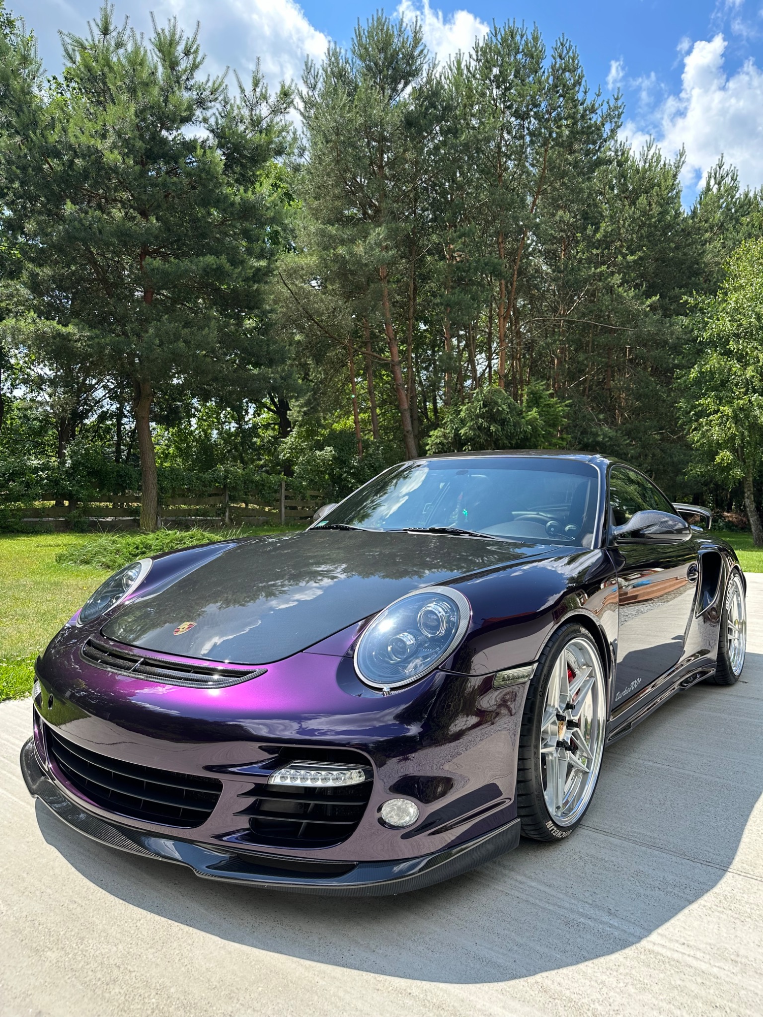 Porsche 911 Turbo w fioletowo-czarnym, opalizującym oklejeniu, zaparkowany na betonowym podjeździe, z lasem w tle.
