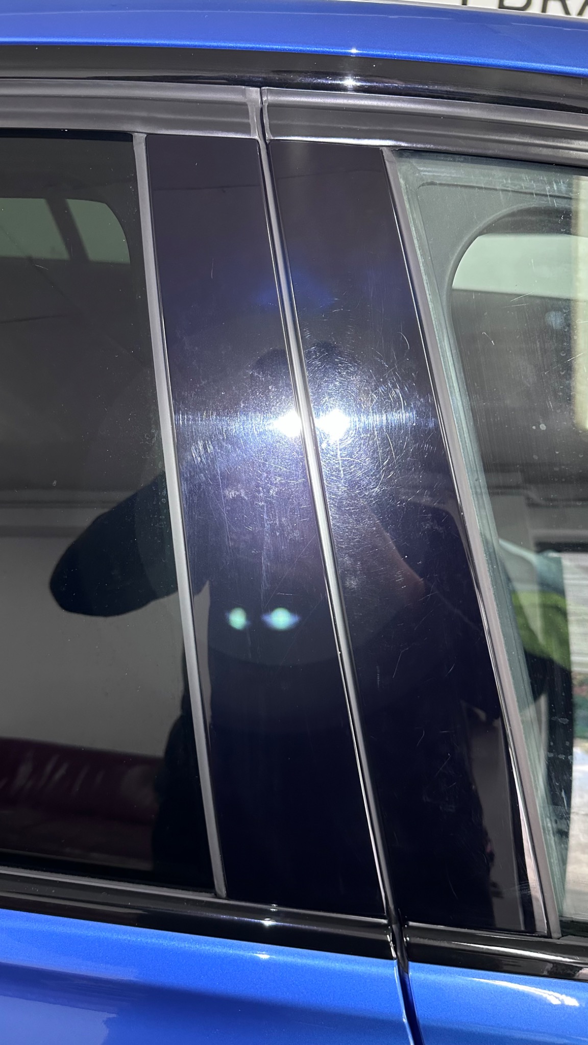 Błyszczący, niebieski samochód z widocznymi zarysowaniami na czarnych, błyszczących słupkach okiennych, odbijające się światło.