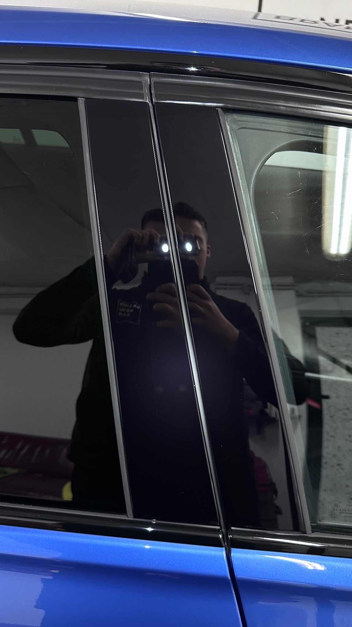 Błyszczące, czarne słupki okienne w niebieskim samochodzie, odbicie fotografa z lampą błyskową w polerowanej powierzchni.