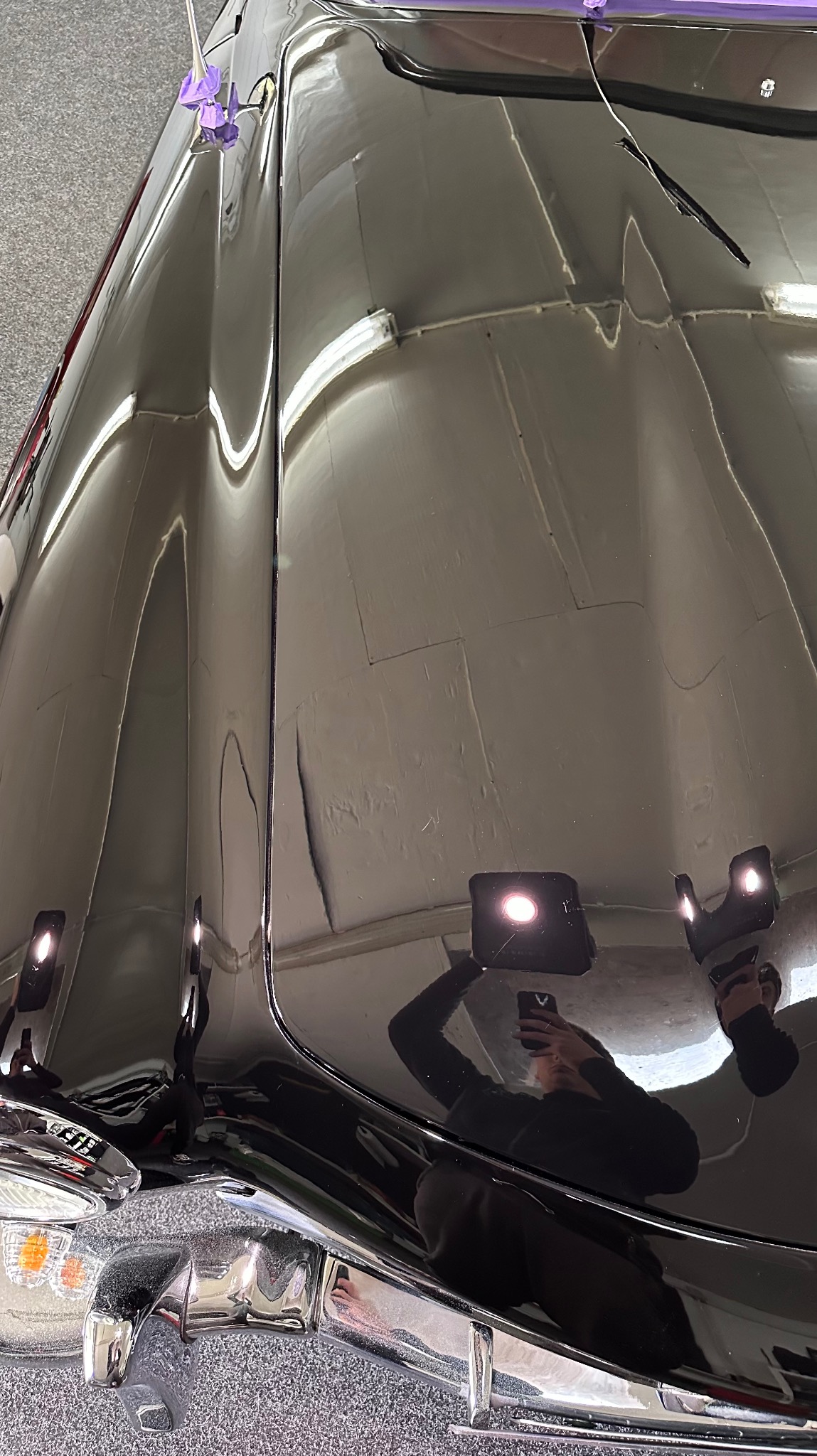 Błyszcząca, czarna karoseria samochodu po polerowaniu, z widocznym odbiciem osoby robiącej zdjęcie telefonem.