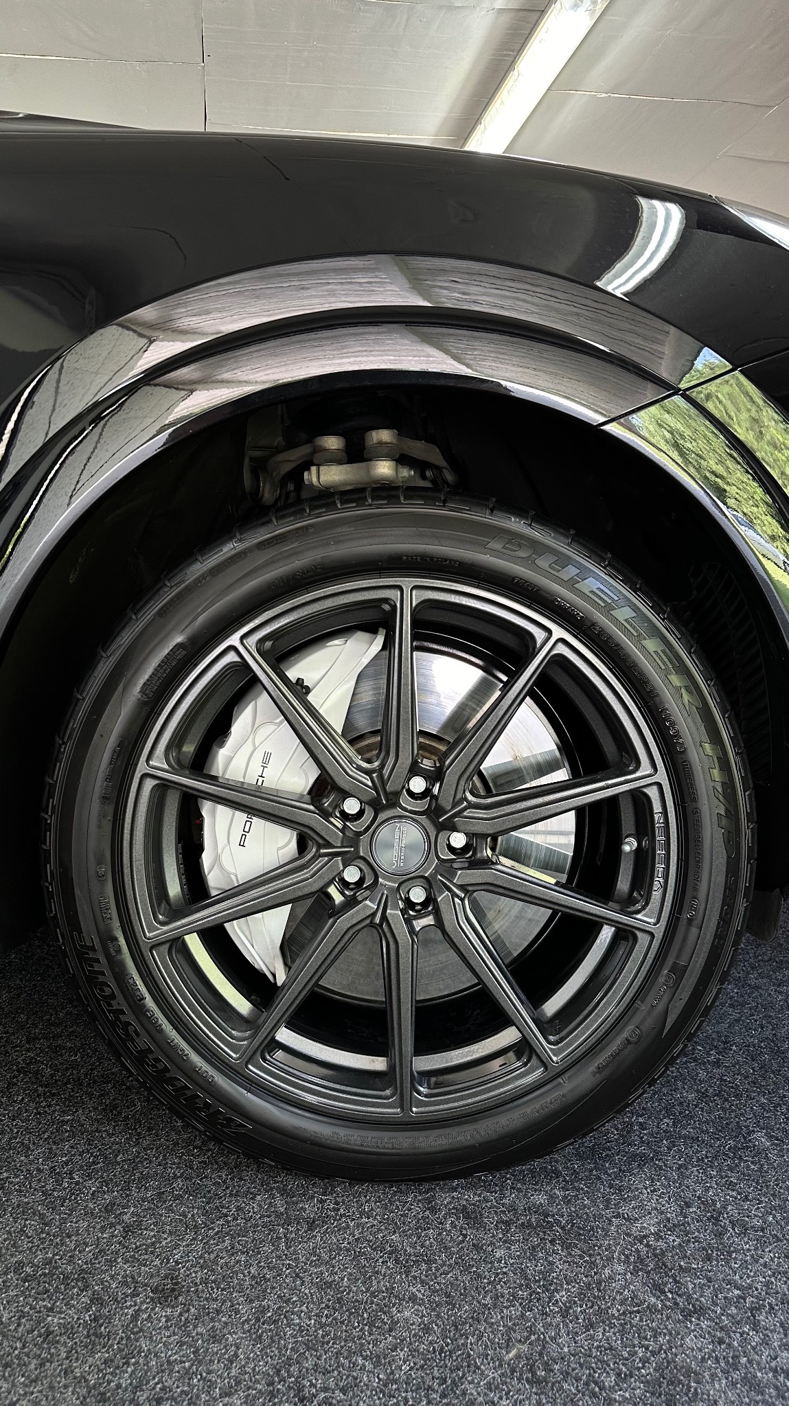 Błyszczące, czarne felgi aluminiowe z widocznym zaciskiem hamulcowym Porsche, opona Bridgestone, czarny lakier samochodu odbijający światło, na tle szarej wykładziny.