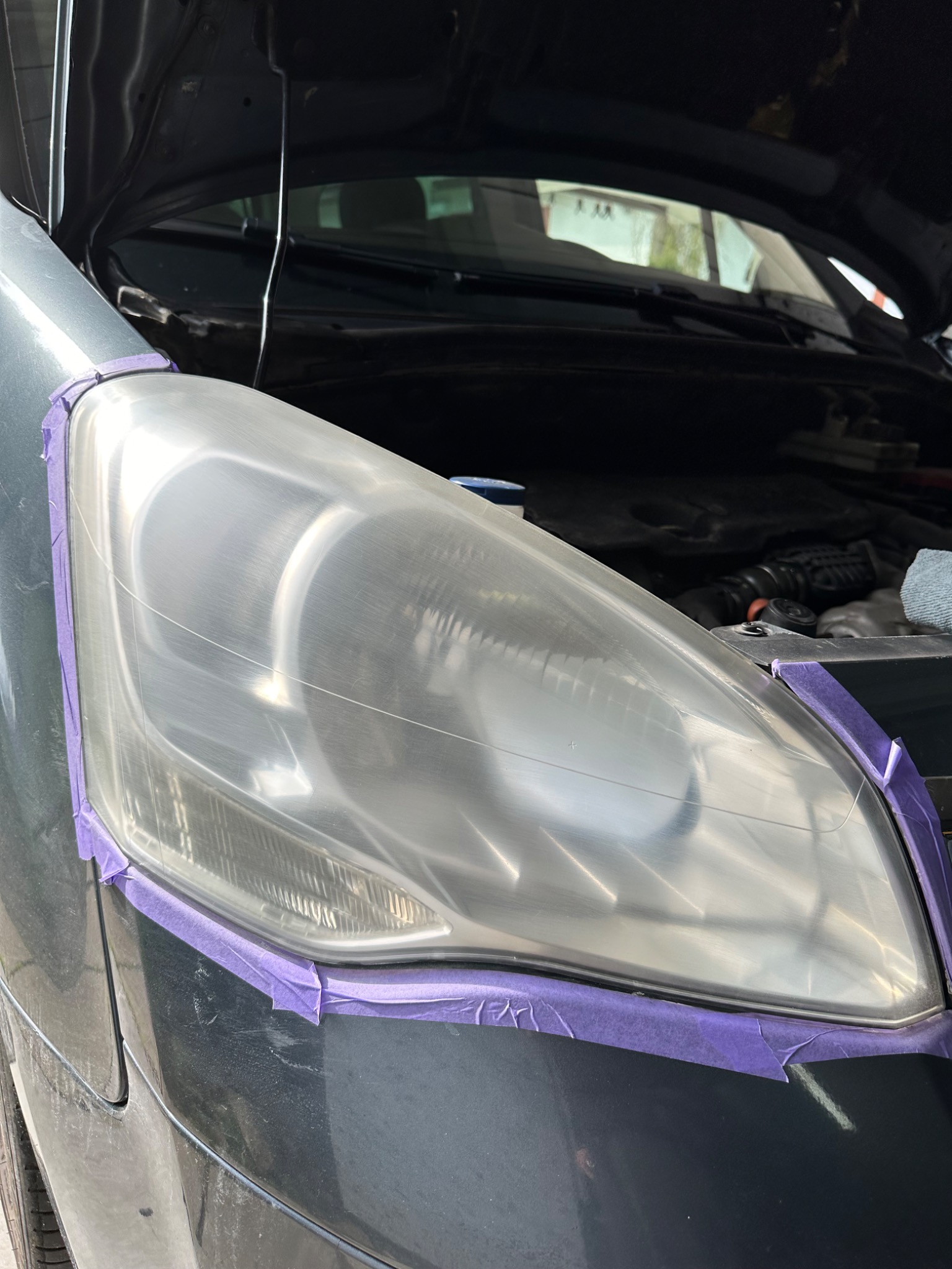 Prawy reflektor samochodu przed renowacją, zabezpieczony fioletową taśmą malarską, z otwartą maską w tle.