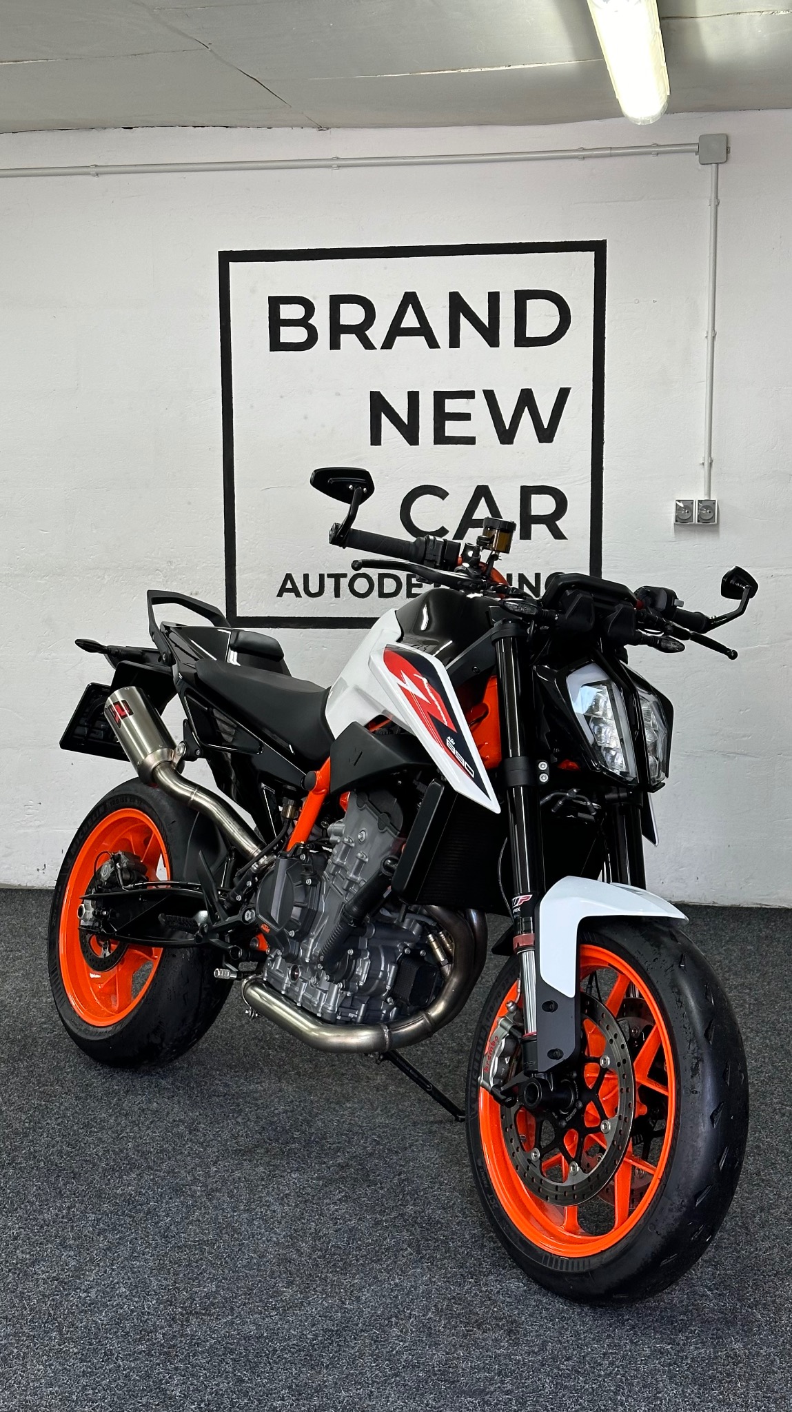 Nowoczesny motocykl KTM z pomarańczowymi felgami na tle białej ściany z czarnym napisem 'BRAND NEW CAR AUTODET'