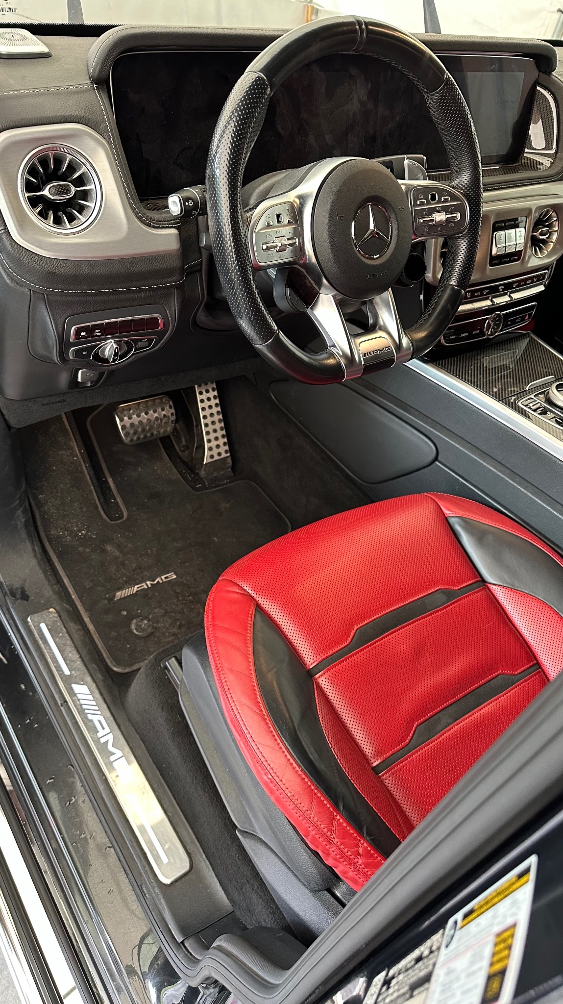 Wnętrze luksusowego samochodu marki Mercedes-Benz AMG z czerwono-czarną skórzaną tapicerką fotela, widoczne detale deski rozdzielczej, kierownica i dywaniki z logo AMG.