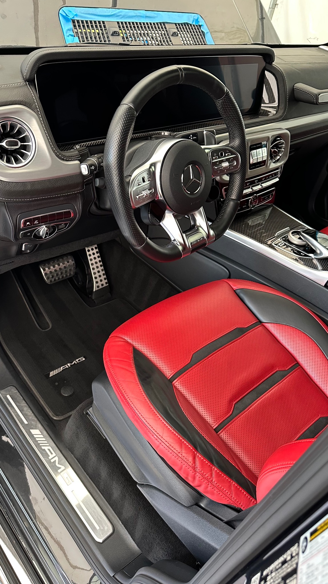 Wnętrze luksusowego samochodu marki Mercedes-Benz AMG z czerwoną perforowaną skórzaną tapicerką fotela kierowcy, czarną kierownicą i dywanikami z logo AMG, widok z boku.