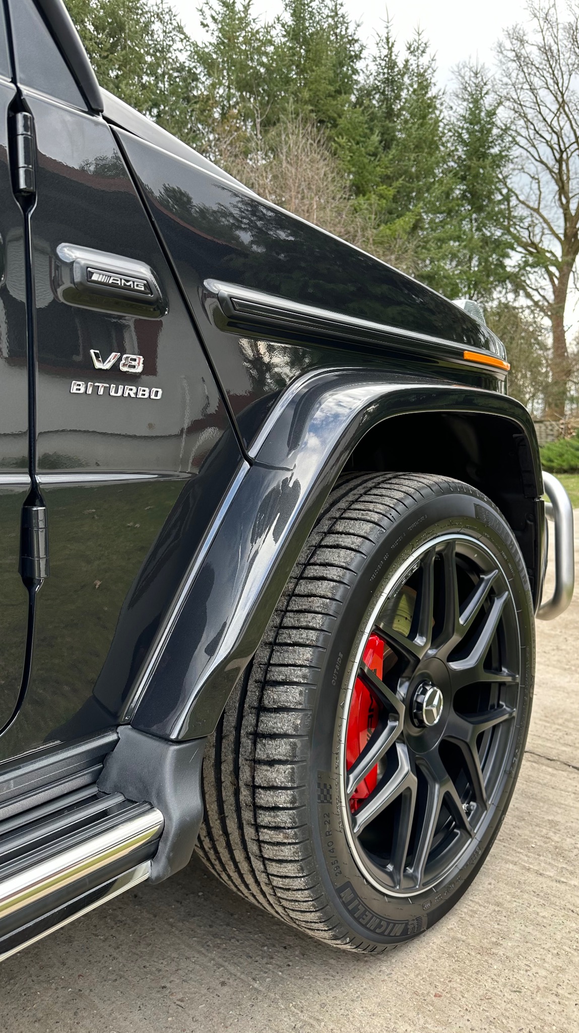 Częściowy widok boku czarnego samochodu marki Mercedes-Benz G-Klasy AMG z oznaczeniami V8 Biturbo, czarnymi felgami i czerwonymi zaciskami hamulcowymi na tle zieleni.