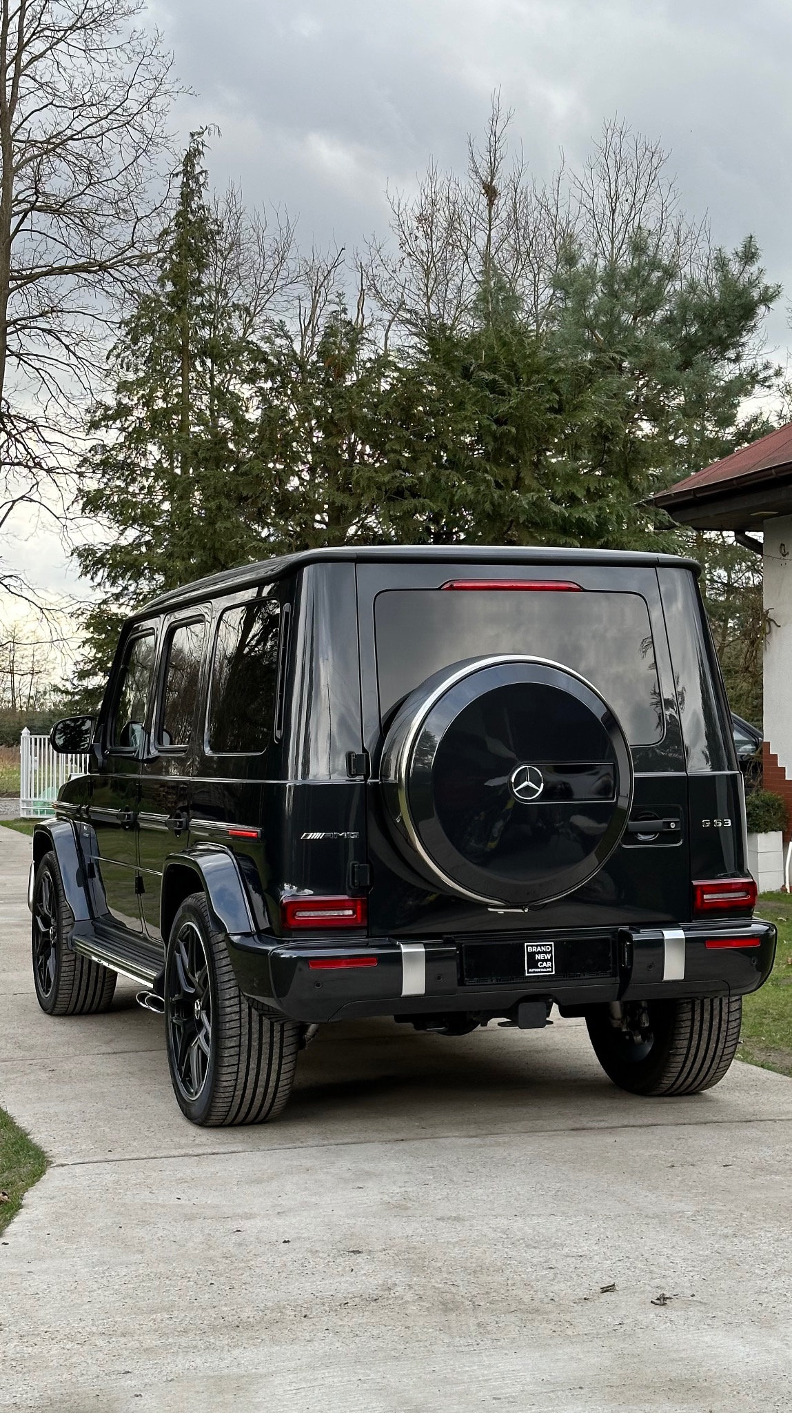 Czarny Mercedes-Benz G 63 AMG z kołem zapasowym na tylnej klapie, widok z tyłu na tle domu i drzew.