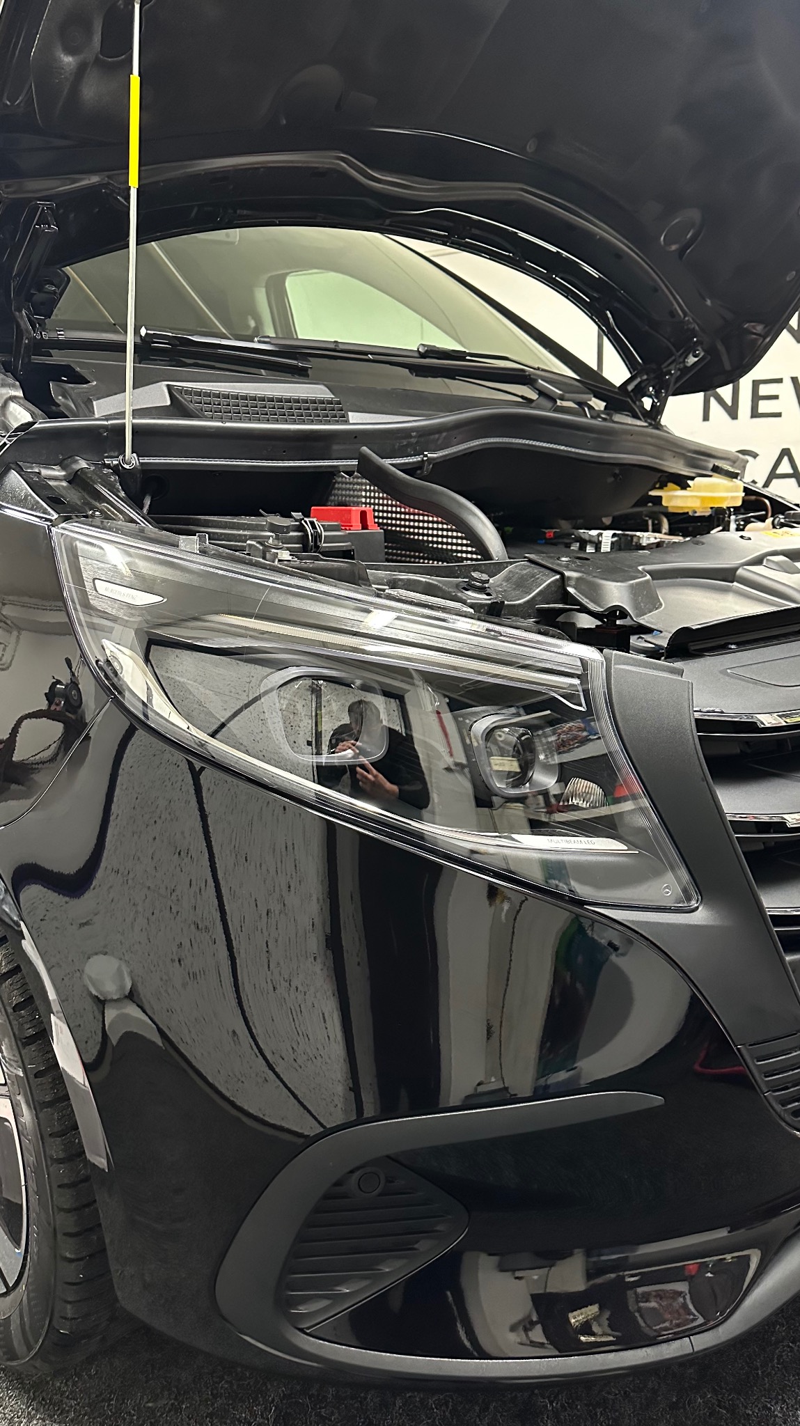 Czarny samochód z otwartą maską, widoczne elementy silnika i odbicie osoby robiącej zdjęcie w reflektorze