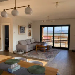 Jasny salon z beżową sofą narożną, drewnianym stolikiem kawowym na wzorzystym dywanie, widok na taras przez duże okno, ceglana ściana w tle.