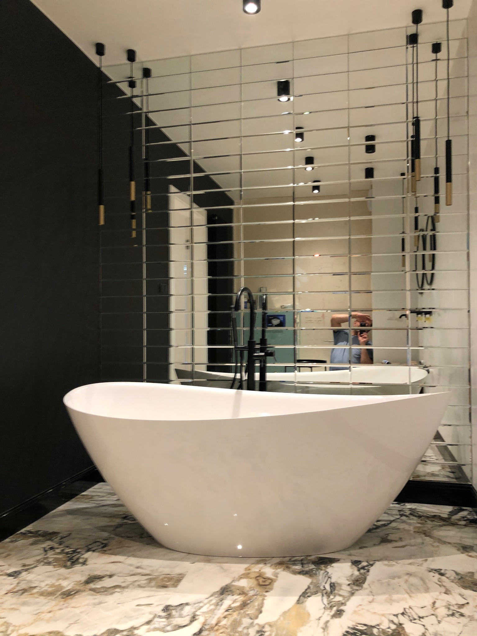Nowoczesna łazienka z wolnostojącą białą wanną na marmurowej posadzce, ściana z lustrzanych płytek i czarne, wiszące lampy.