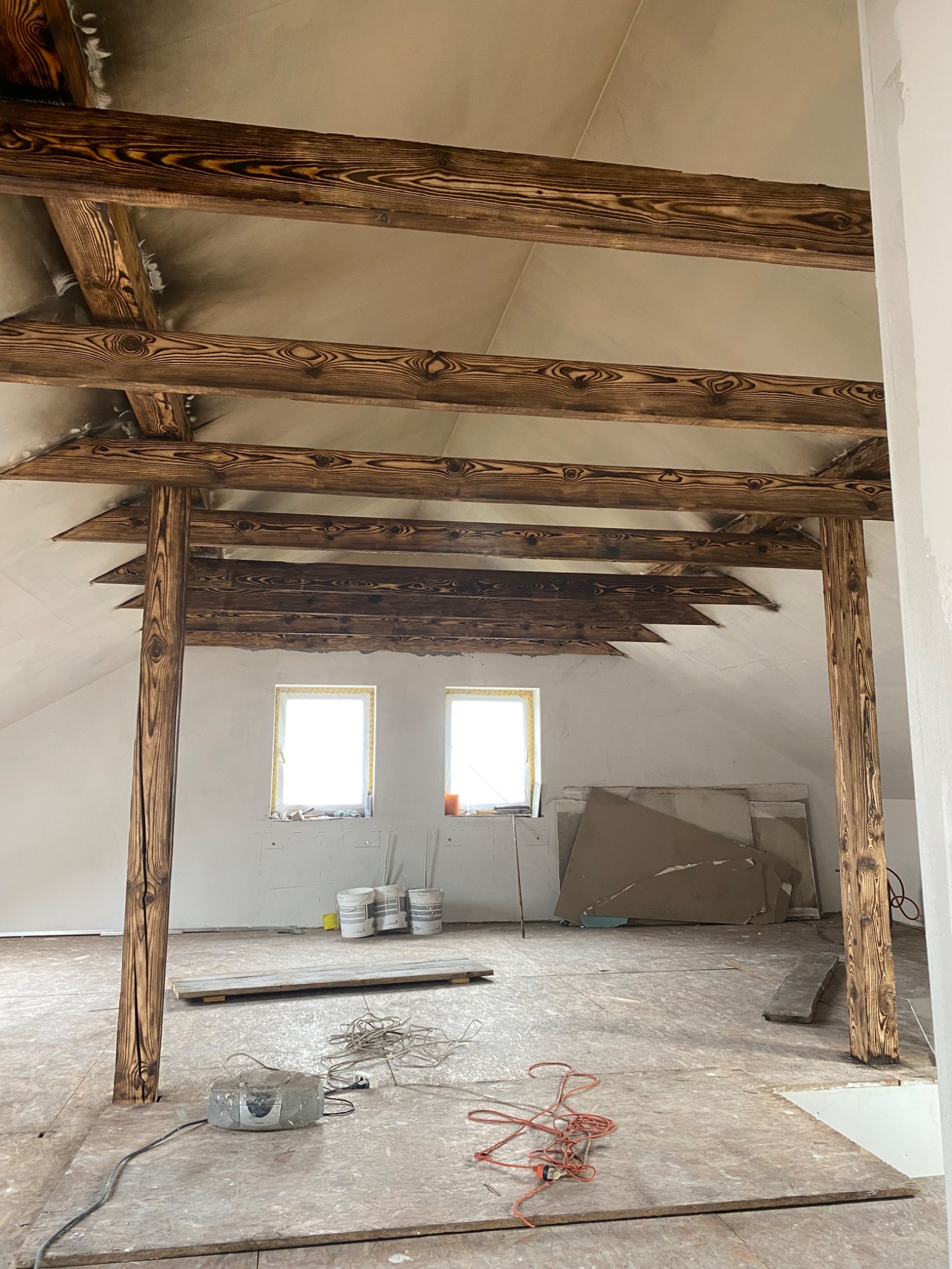 Poddasze w trakcie prac wykończeniowych, widoczne drewniane belki stropowe o wyraźnym usłojeniu, surowe ściany, płyty karton-gips, wiadra z farbą i narzędzia na podłodze.