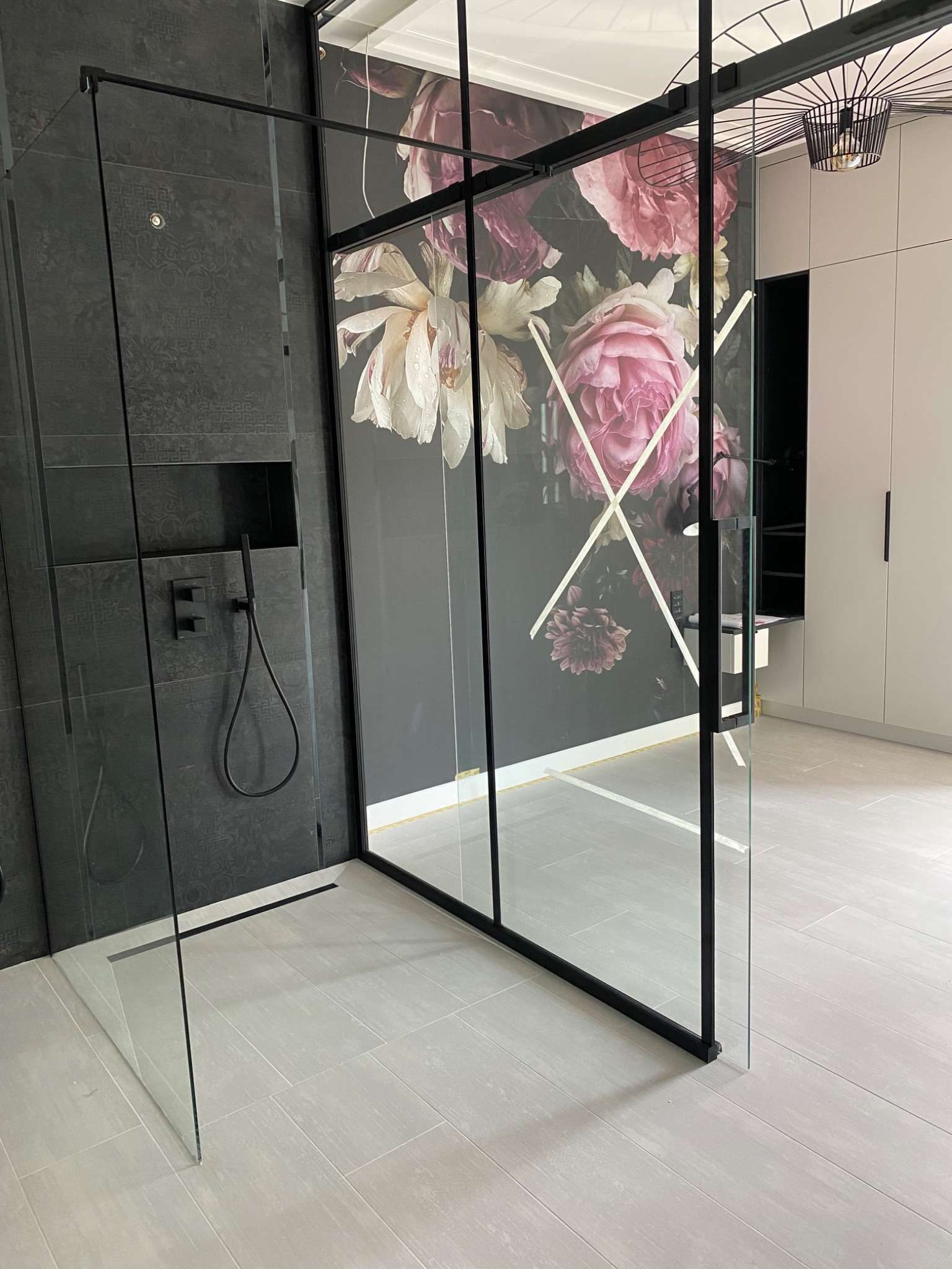 Nowoczesna kabina prysznicowa walk-in z czarnymi profilami i szkłem, z czarną armaturą na ścianie z ciemnoszarych płytek z subtelnym wzorem, ozdobiona fototapetą z motywem kwiatowym w odcieniach...