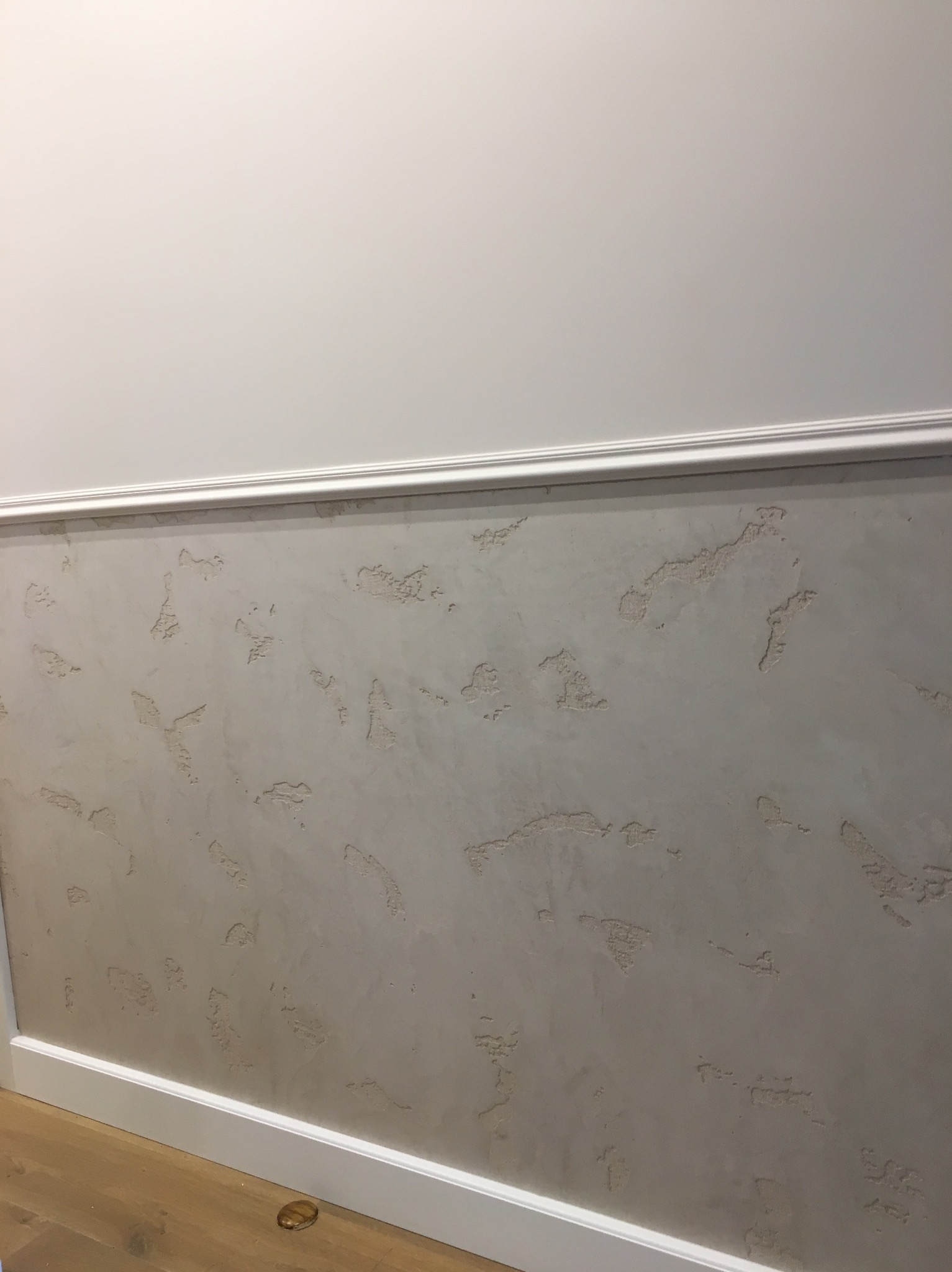 Fragment ściany z dekoracyjną fakturą szpachlową w odcieniach szarości, oddzielonej listwą od gładkiej, białej części górnej, widoczna drewniana podłoga.