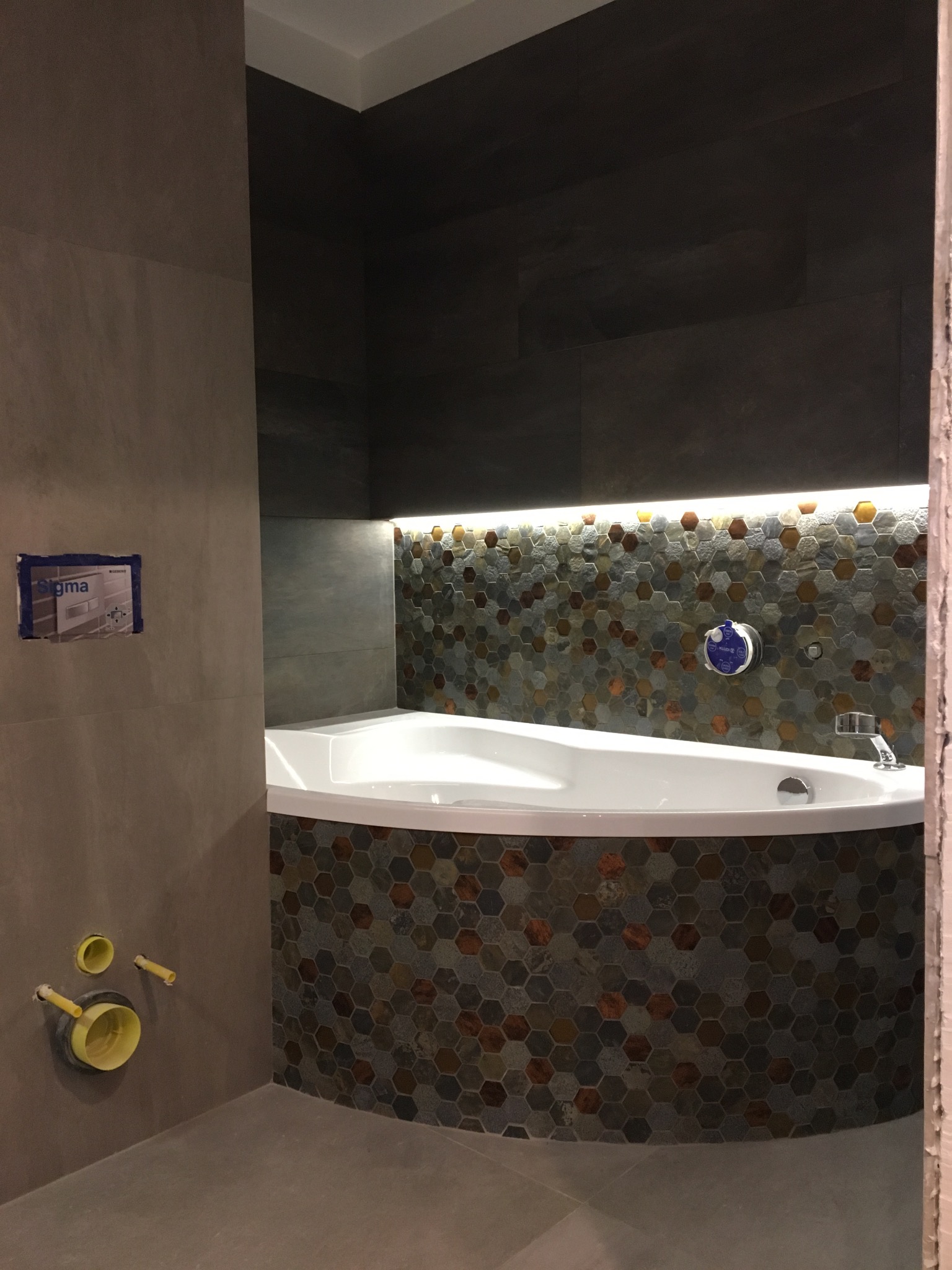 Narożna wanna obudowana mozaiką heksagonalną w odcieniach szarości i brązu, podświetlona listwą LED, z widoczną instalacją sanitarną w ścianie i częściowo zamaskowanym stelażem podtynkowym.