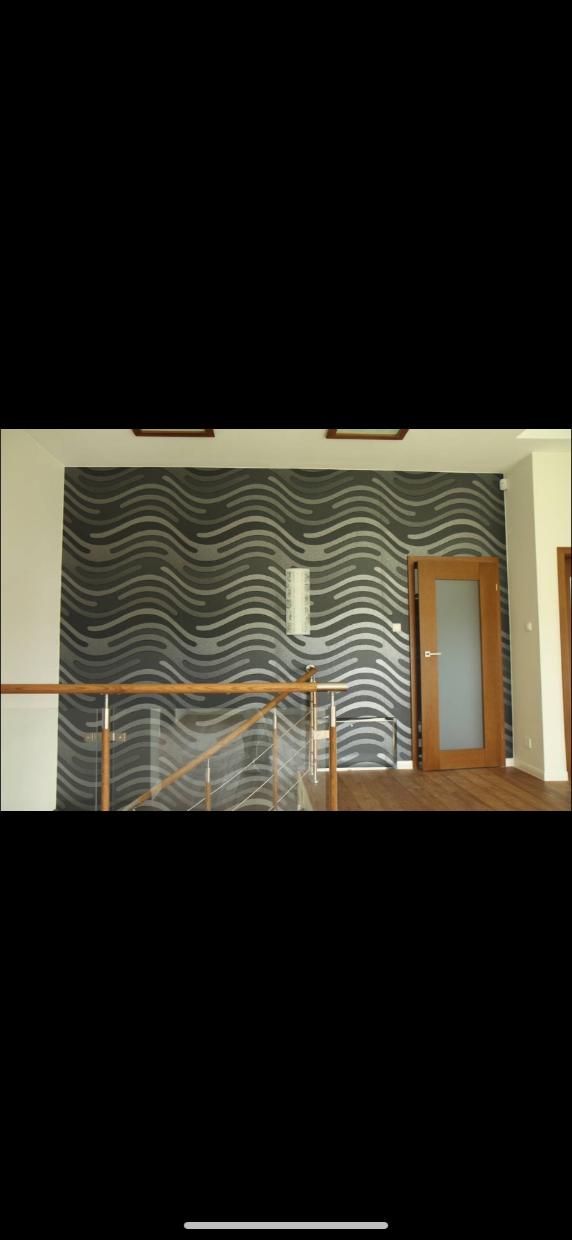 Ściana z tapetą w szare fale, drewniane drzwi i podłoga, widok zza szklanej balustrady.
