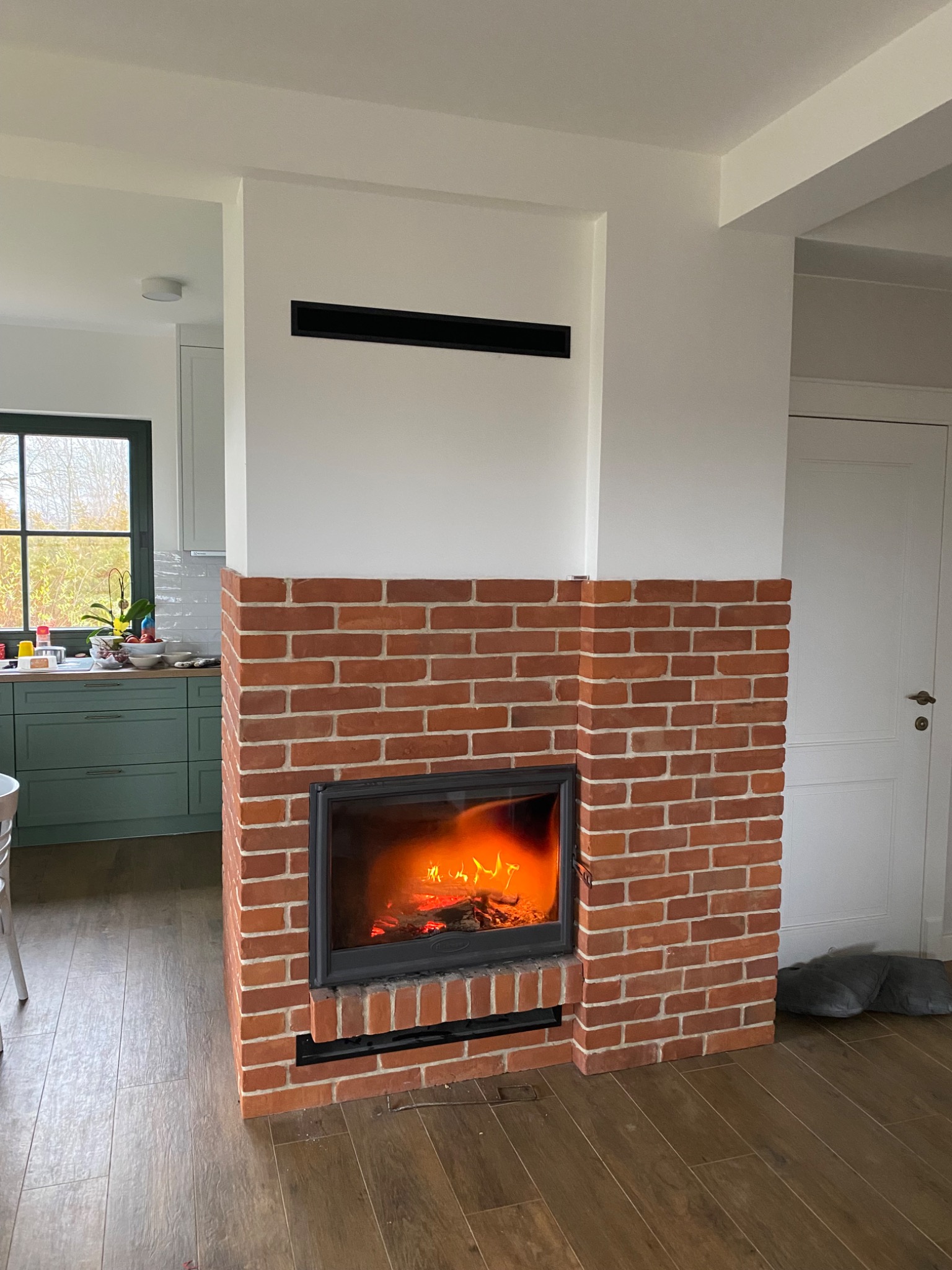 Nowoczesny kominek obudowany czerwoną cegłą w jasnym wnętrzu, widoczne płomienie, drewniana podłoga i fragment kuchni w tle.