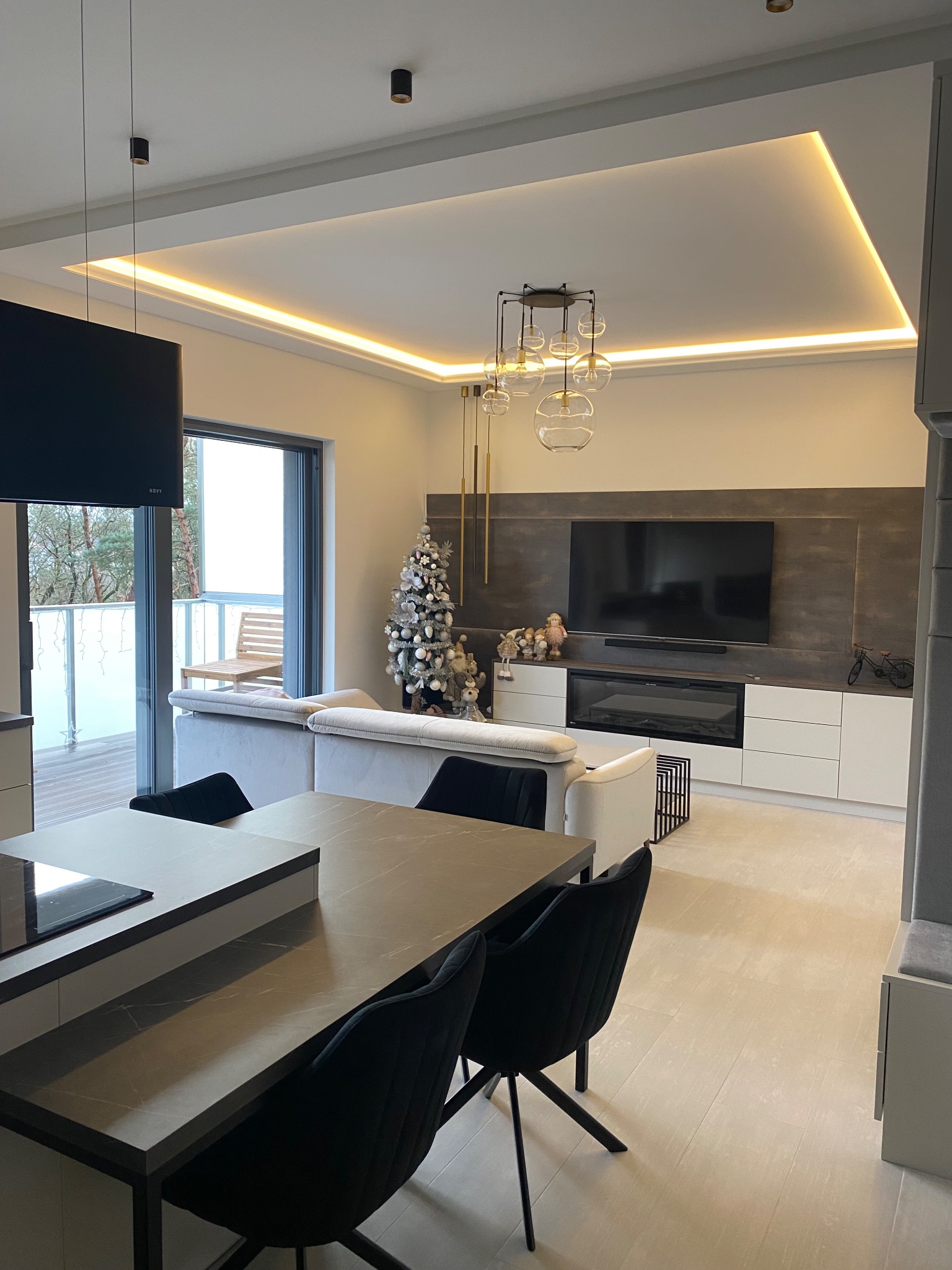 Nowoczesny salon z aneksem kuchennym w szarościach, z podwieszanym sufitem z oświetleniem LED, szarą ścianą TV, świątecznym drzewkiem i wyjściem na balkon.