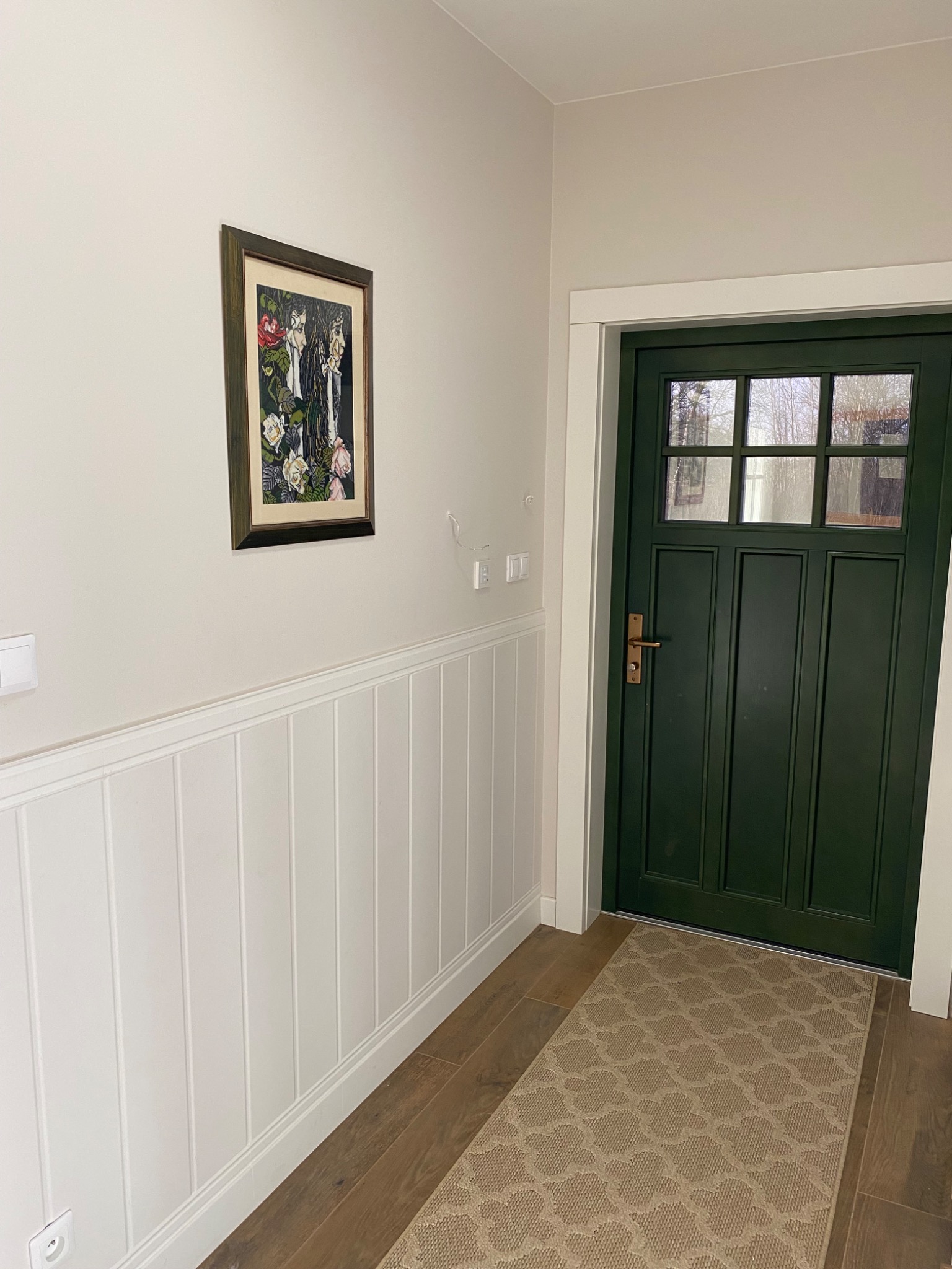 Korytarz w Olsztynie z ciemnozielonymi drzwiami wejściowymi i boazerią angielską pomalowaną na biało, na ścianie wisi obraz w ozdobnej ramie, a na podłodze leży beżowy dywanik.