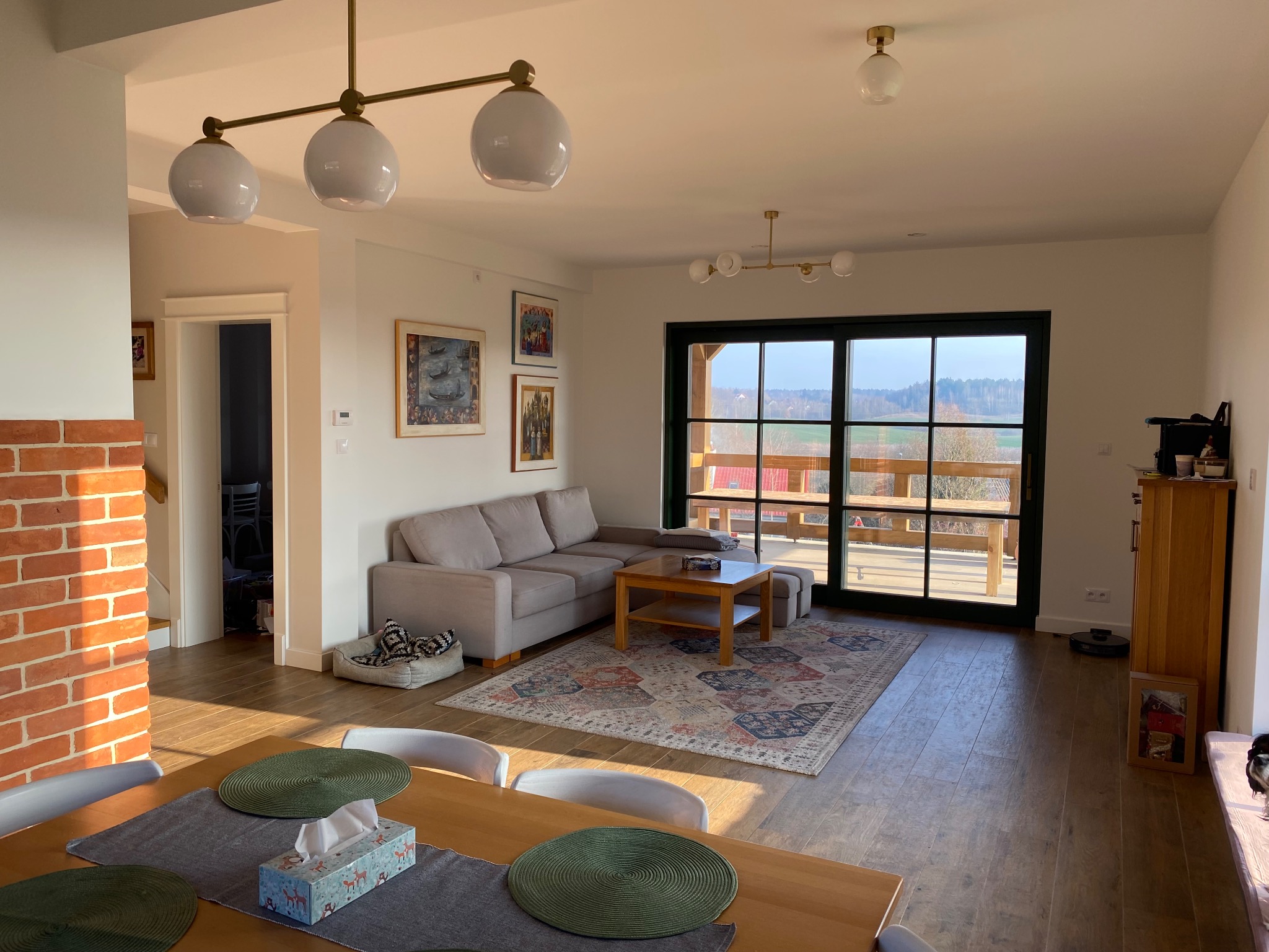 Jasny salon z beżową sofą narożną, drewnianym stolikiem kawowym na wzorzystym dywanie, widok na taras przez duże okno, ceglana ściana w tle.