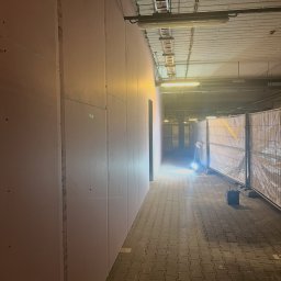 Simbud - Korytarz w trakcie remontu, ściany wyłożone płytami kartonowo-gipsowymi, widoczne zabezpieczenia i oświetlenie budowlane. Prace wykończeniowe w przestrzeni komercyjnej.