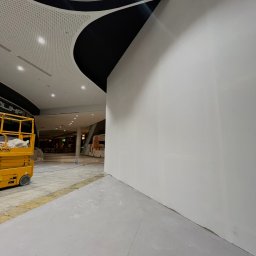 Simbud - Biała, gładka ściana w centrum handlowym po szpachlowaniu, z widocznym podnośnikiem i elementami sufitu podwieszanego. Ujęcie z dołu, perspektywa zbieżna.