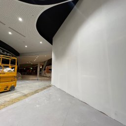 Simbud - Biała, świeżo pomalowana ściana w galerii handlowej, widoczny podnośnik koszowy. Sufit z perforowanej blachy z czarnym, dekoracyjnym elementem.