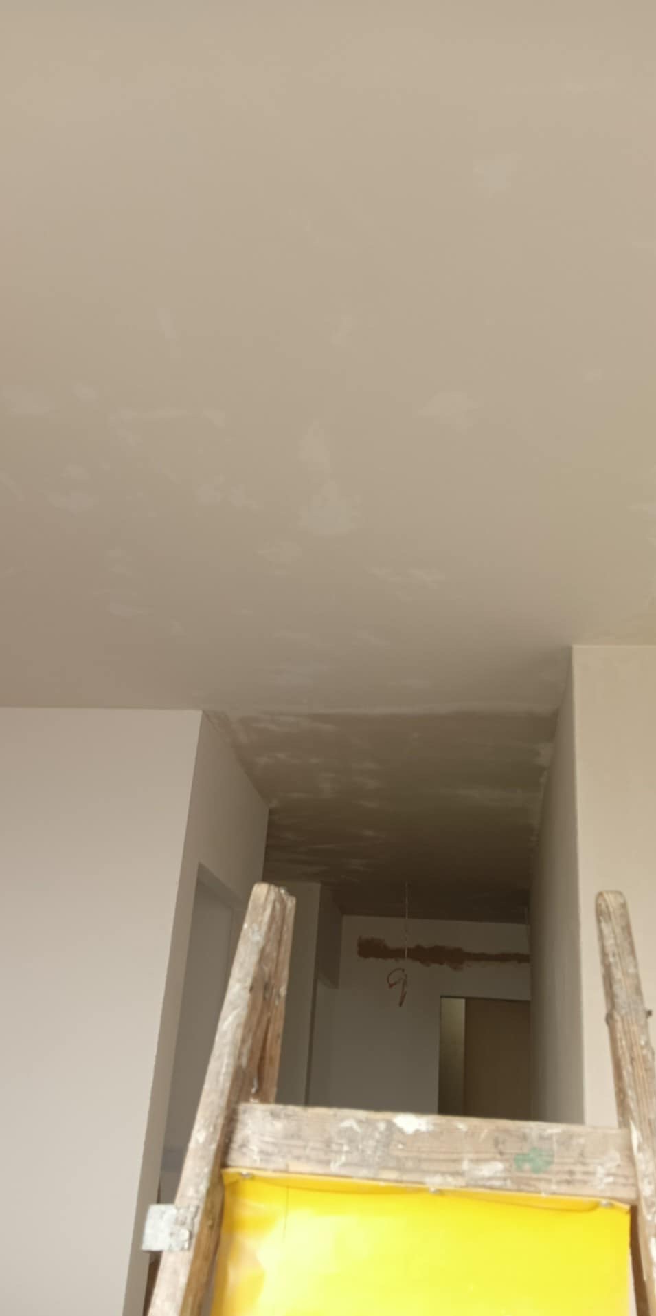 Widok z drabiny na sufit i fragment korytarza w trakcie wykańczania, widoczne ślady po szpachlowaniu i przygotowaniu do malowania. Wiszące przewody elektryczne.