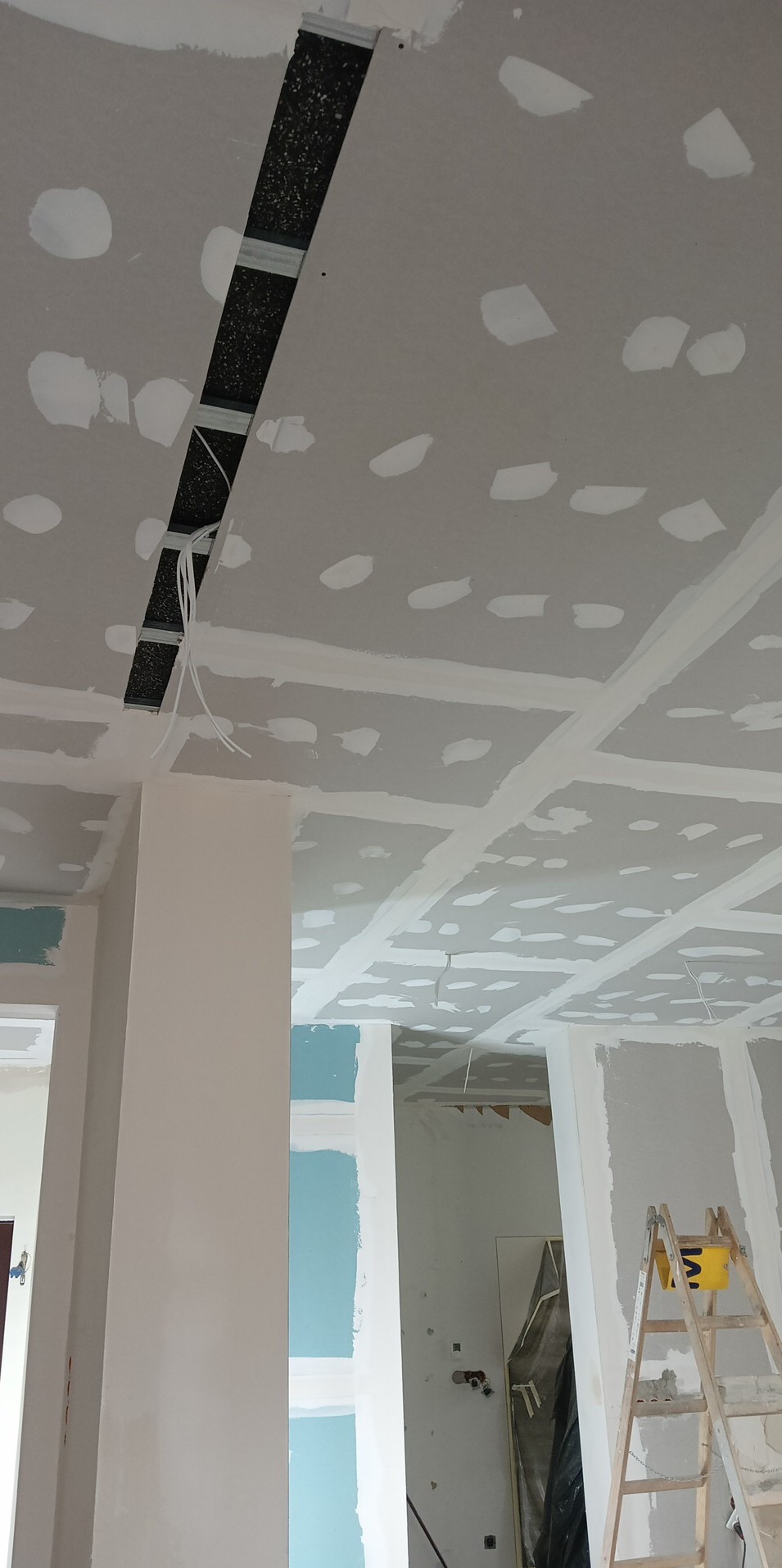 Sufit z płyt kartonowo-gipsowych w trakcie prac wykończeniowych, widoczne łączenia płyt zabezpieczone taśmą i szpachlą, otwór z widoczną instalacją, drabina oparta o ścianę w tle.