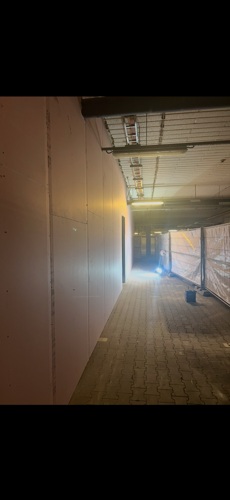 Korytarz w trakcie remontu, ściany wyłożone płytami kartonowo-gipsowymi, widoczne zabezpieczenia i oświetlenie budowlane. Prace wykończeniowe w przestrzeni komercyjnej.