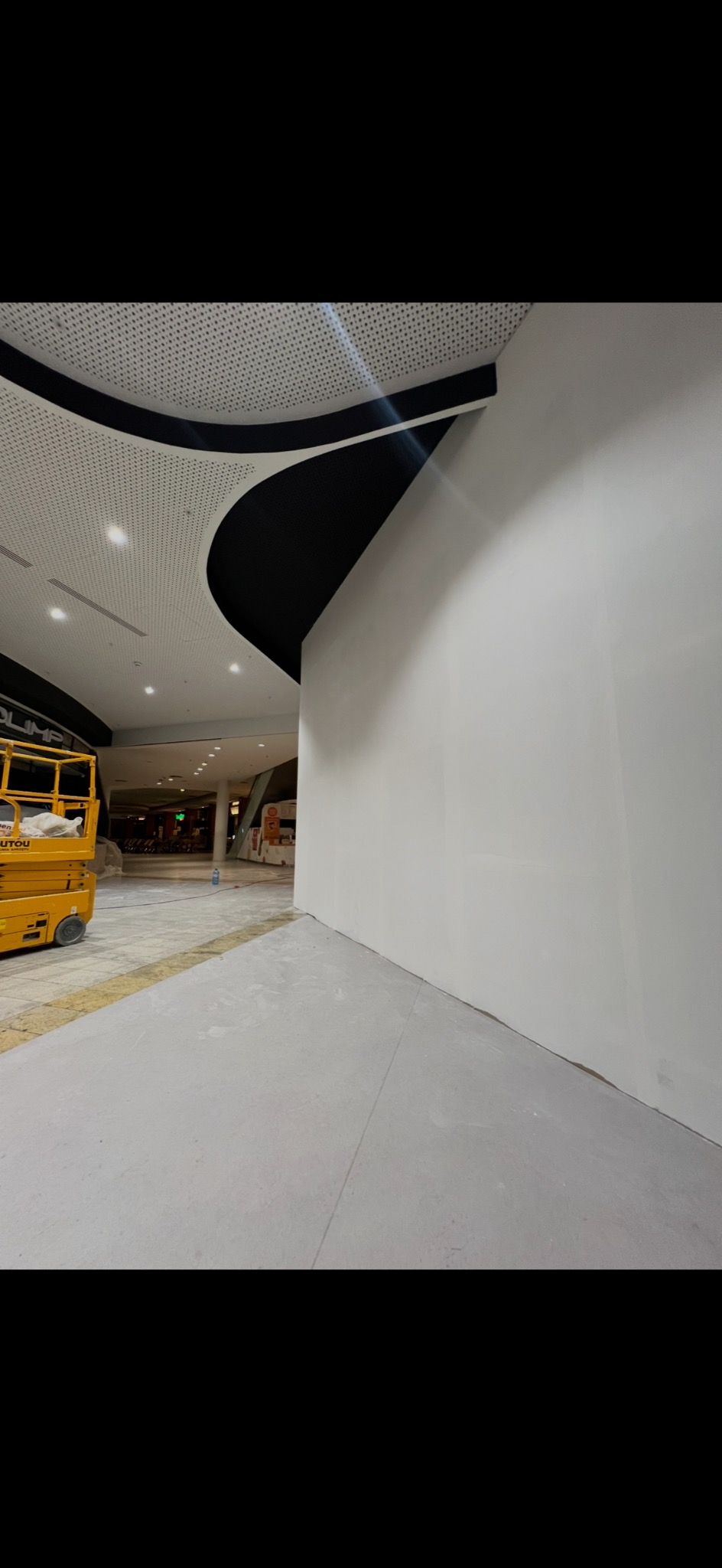 Biała, gładka ściana w centrum handlowym po szpachlowaniu, z widocznym podnośnikiem i elementami sufitu podwieszanego. Ujęcie z dołu, perspektywa zbieżna.