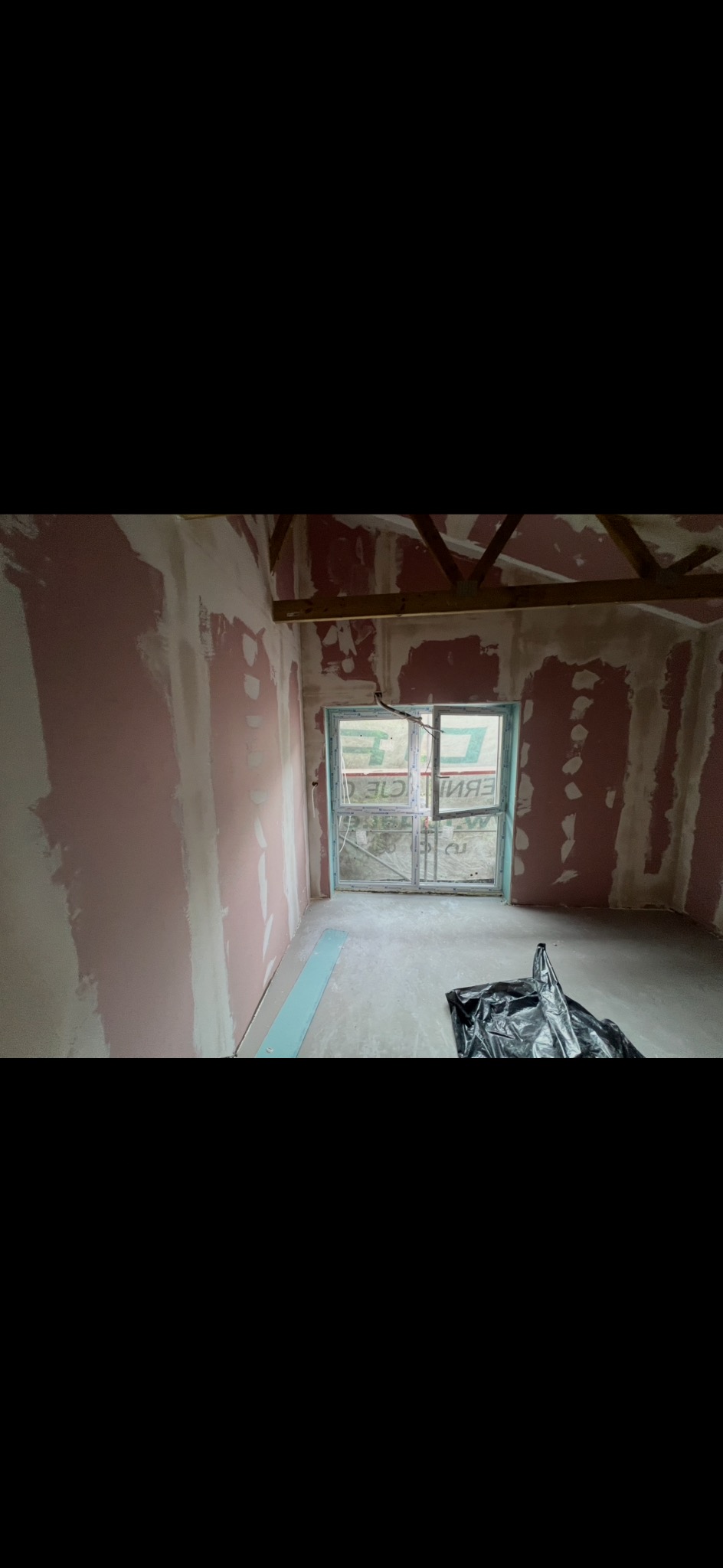 Pokój w trakcie prac wykończeniowych: ściany częściowo pokryte gładzią, widoczne fragmenty płyty karton-gips, okno z folią ochronną, podłoga przygotowana do dalszych prac.