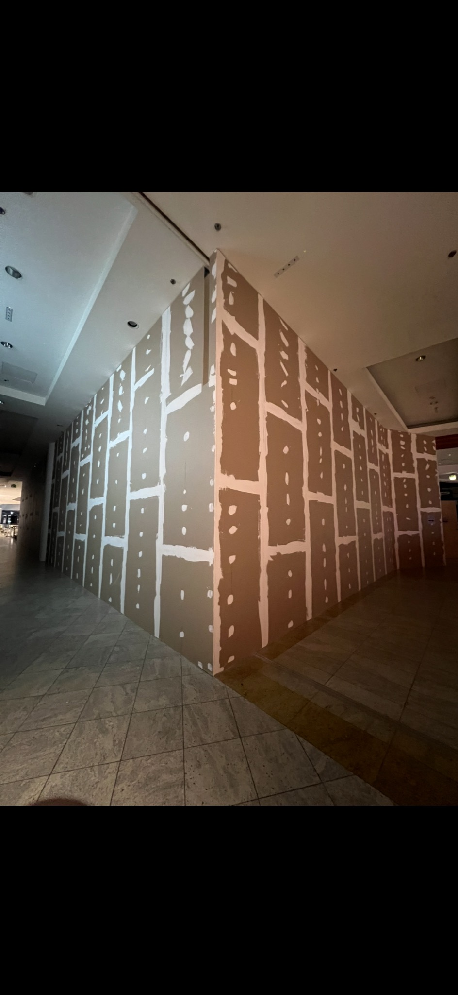 Ściana z płyt kartonowo-gipsowych, z widocznymi łączeniami szpachlowanymi na biało, w przestrzeni komercyjnej z marmurową podłogą i oświetleniem sufitowym.