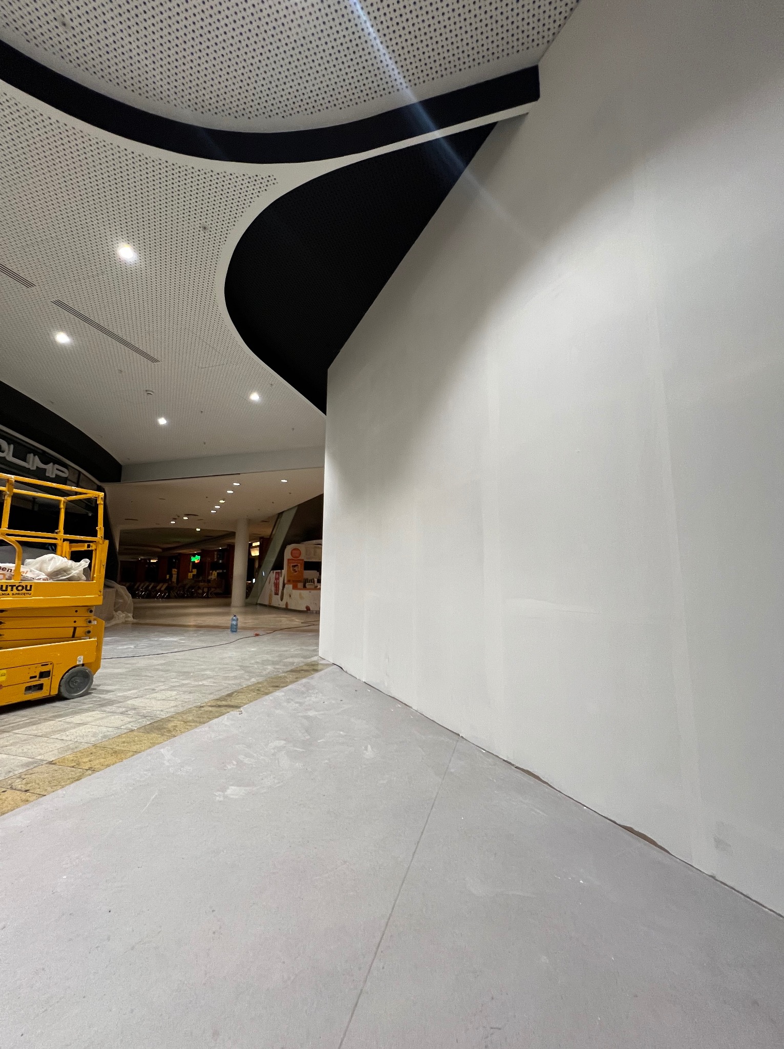 Biała, świeżo pomalowana ściana w galerii handlowej, widoczny podnośnik koszowy. Sufit z perforowanej blachy z czarnym, dekoracyjnym elementem.