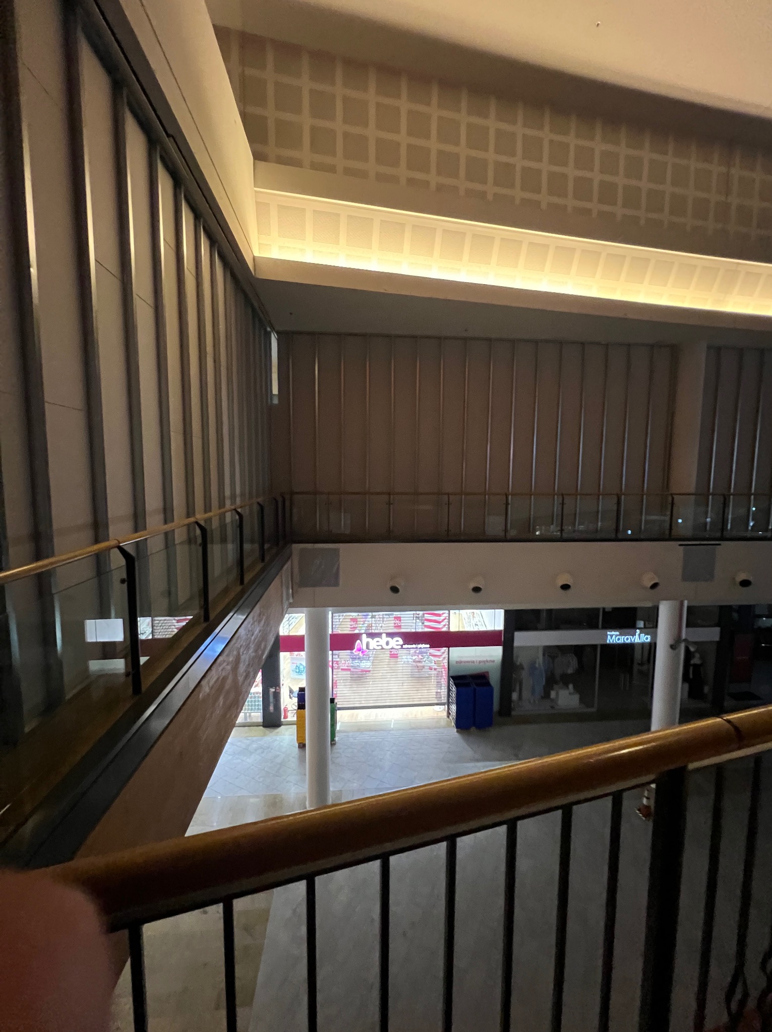 Wnętrze galerii handlowej z widokiem na sklepy z poziomu antresoli. Widoczne balustrady, oświetlenie sufitowe i witryny sklepowe.