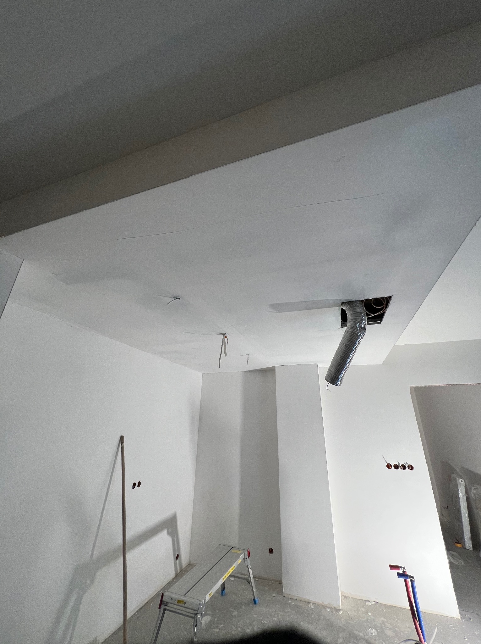 Białe wnętrze w trakcie prac wykończeniowych: sufit podwieszany z otworem wentylacyjnym, drabina aluminiowa, ściany przygotowane do malowania, widoczne instalacje.