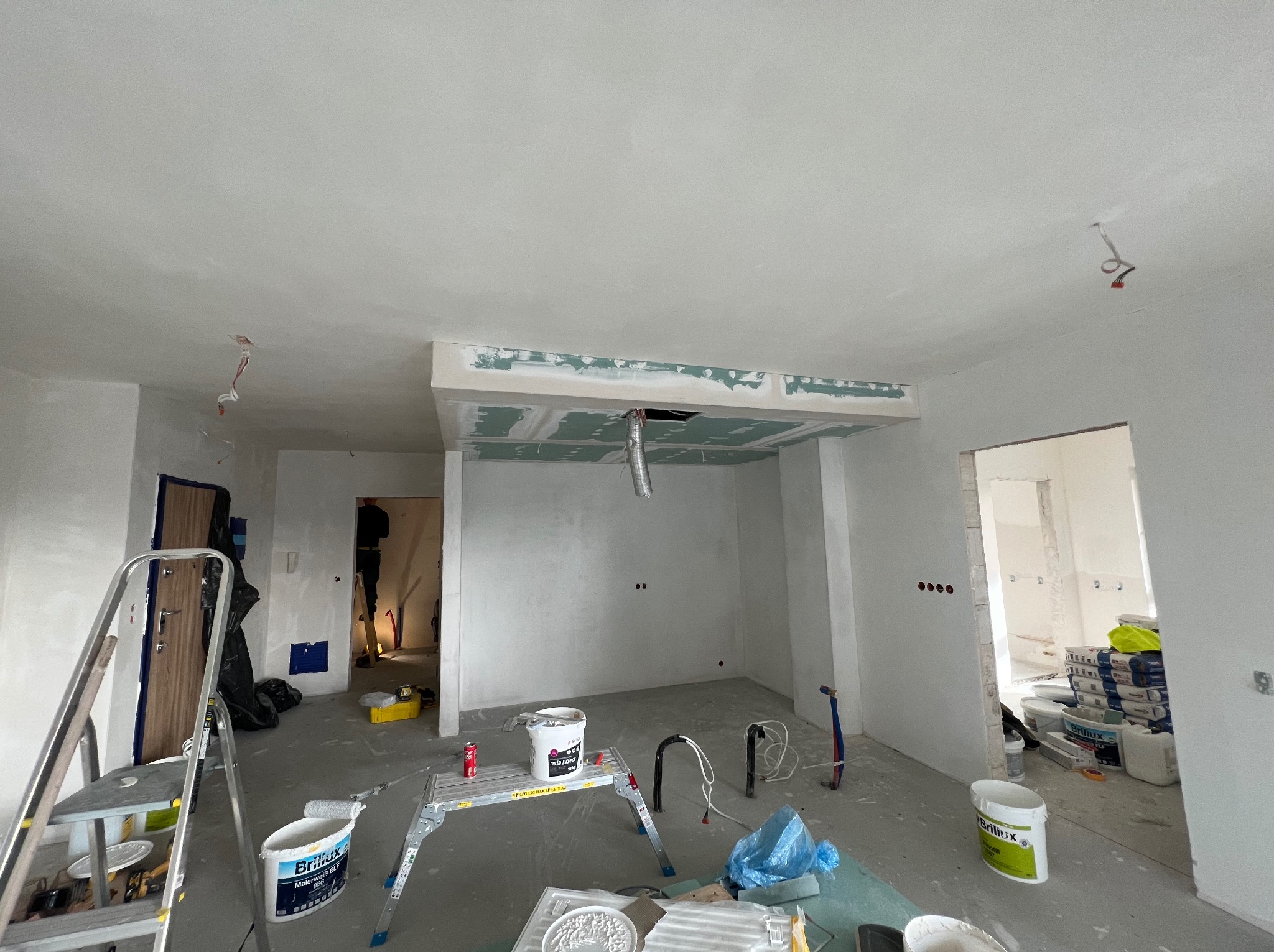 Wnętrze w trakcie prac wykończeniowych: sufit z płyt karton-gips, ściany po szpachlowaniu, drabina, wiadra z farbą. Widoczne kable i instalacje.