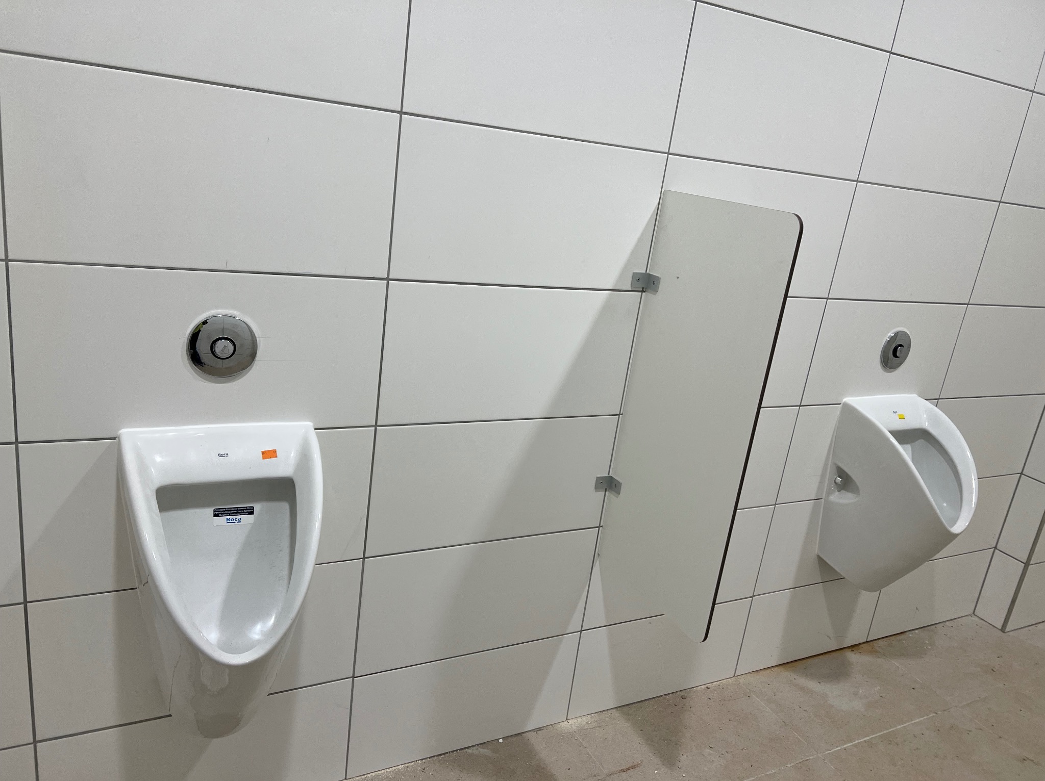 Białe pisuary na ścianie z białych płytek, jeden z przegrodą. Widoczne przyciski spłukiwania i drobne niedoskonałości na podłodze. Minimalistyczny design.