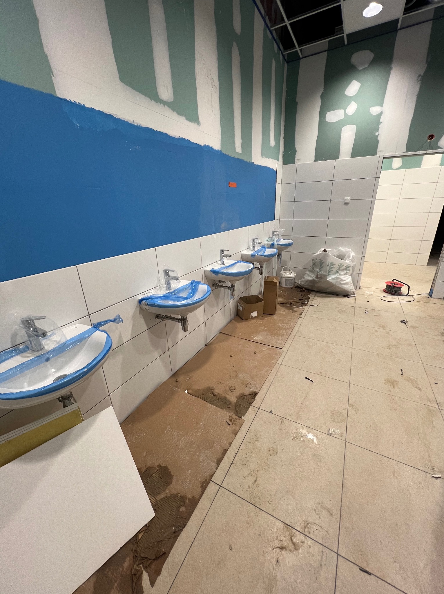 Wnętrze łazienki w trakcie remontu: ściany z płyt gipsowo-kartonowych, pomalowana na niebiesko górna część ściany, białe płytki, umywalki zabezpieczone folią.