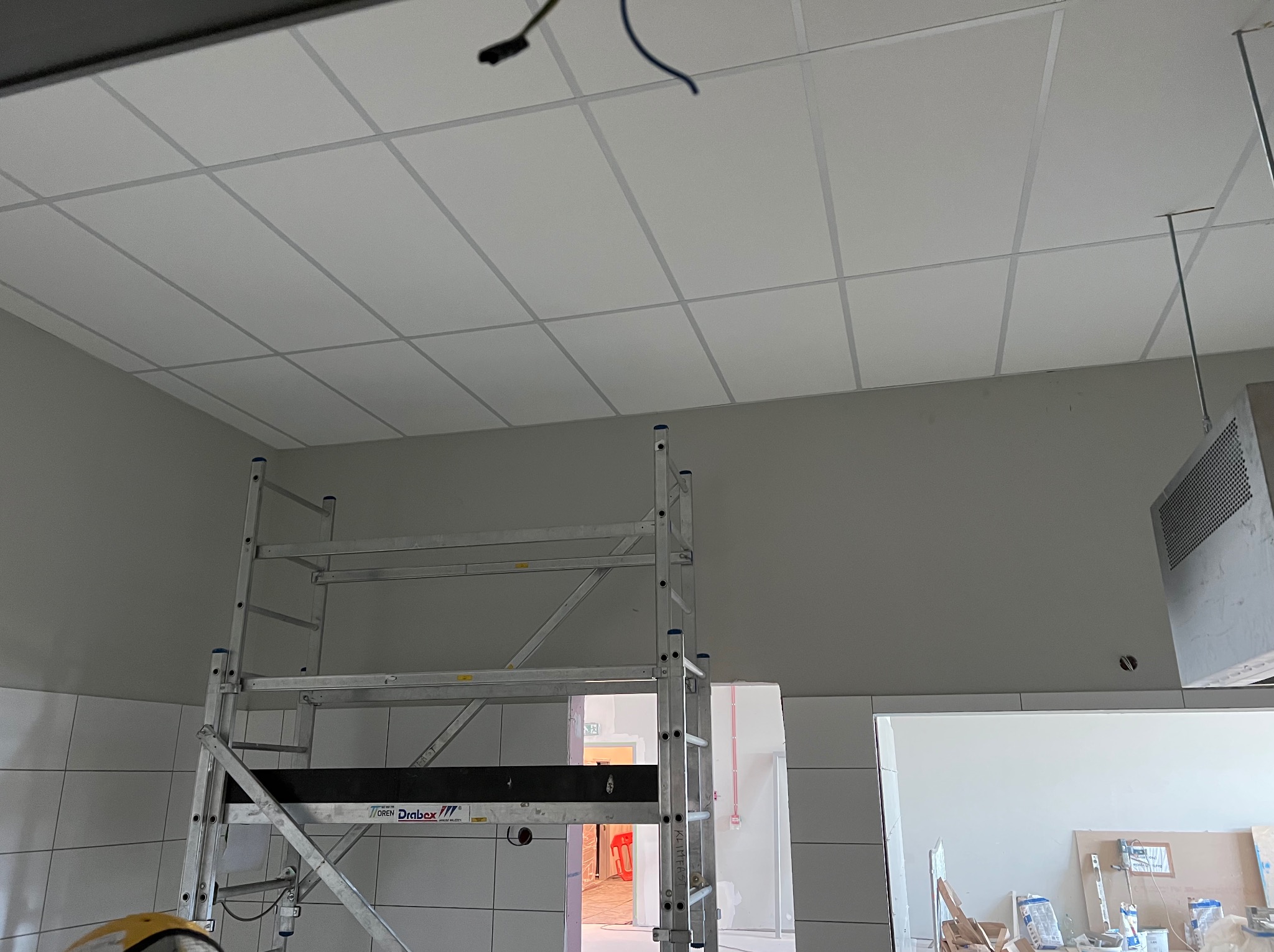 Sufit kasetonowy w trakcie montażu, widoczny z poziomu rusztowania. W tle fragment ściany i otwarte przejście do innego pomieszczenia.