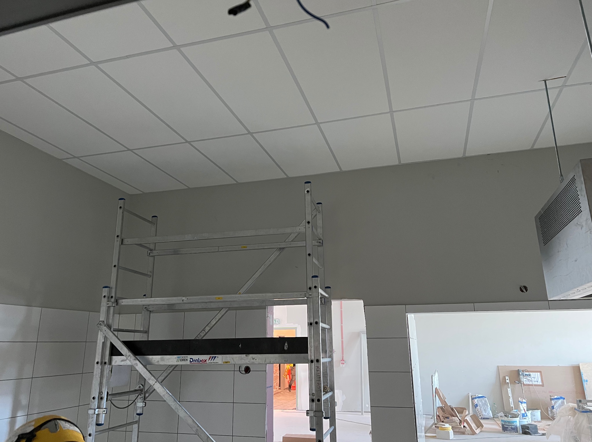 Montaż sufitu podwieszanego kasetonowego w pomieszczeniu z rusztowaniem. Widoczne kafelki na ścianach i materiały budowlane w tle. Prace wykończeniowe.