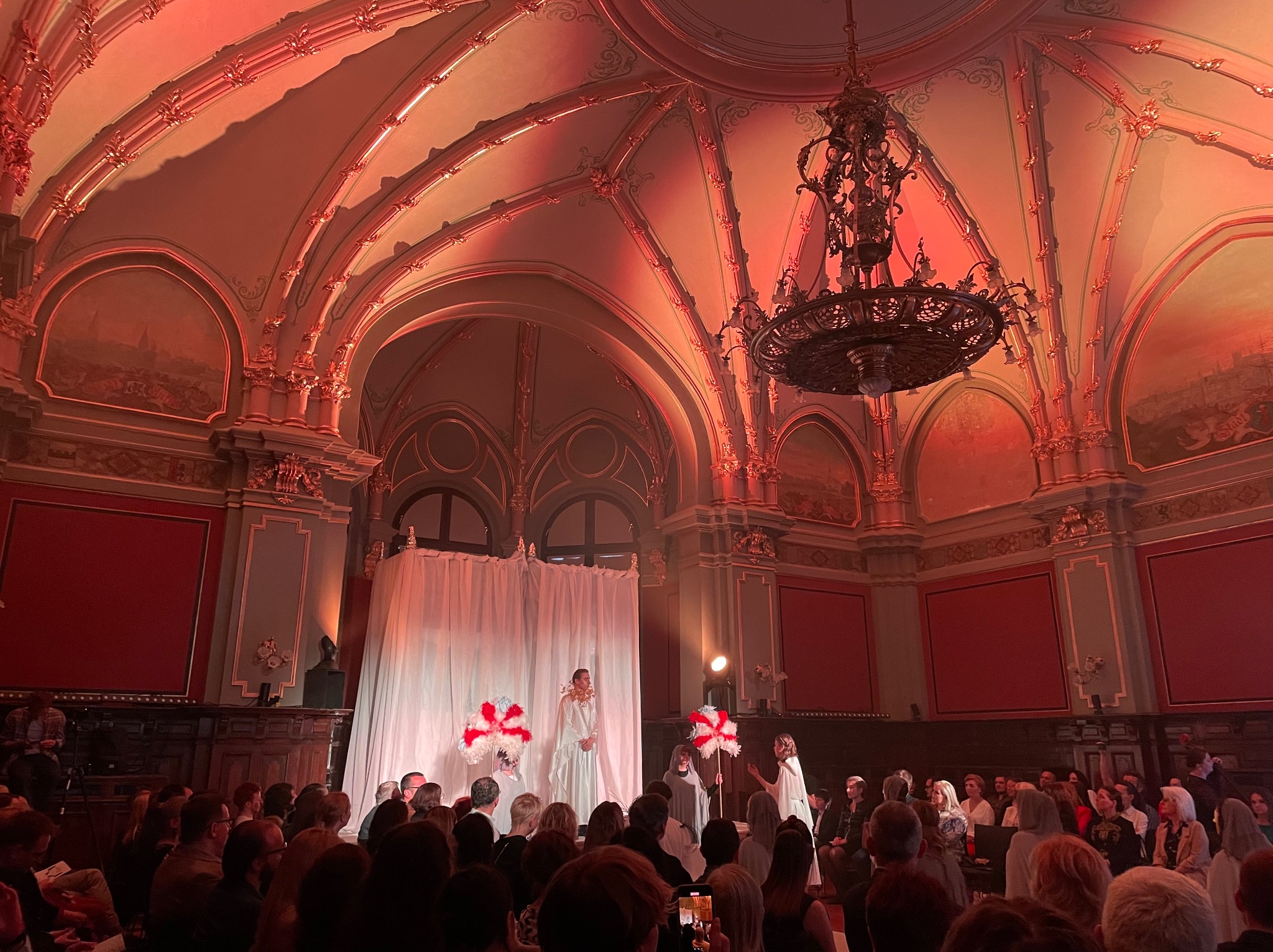 Wnętrze sali balowej oświetlone czerwonym światłem, z widoczną sceną, publicznością i żyrandolem.
